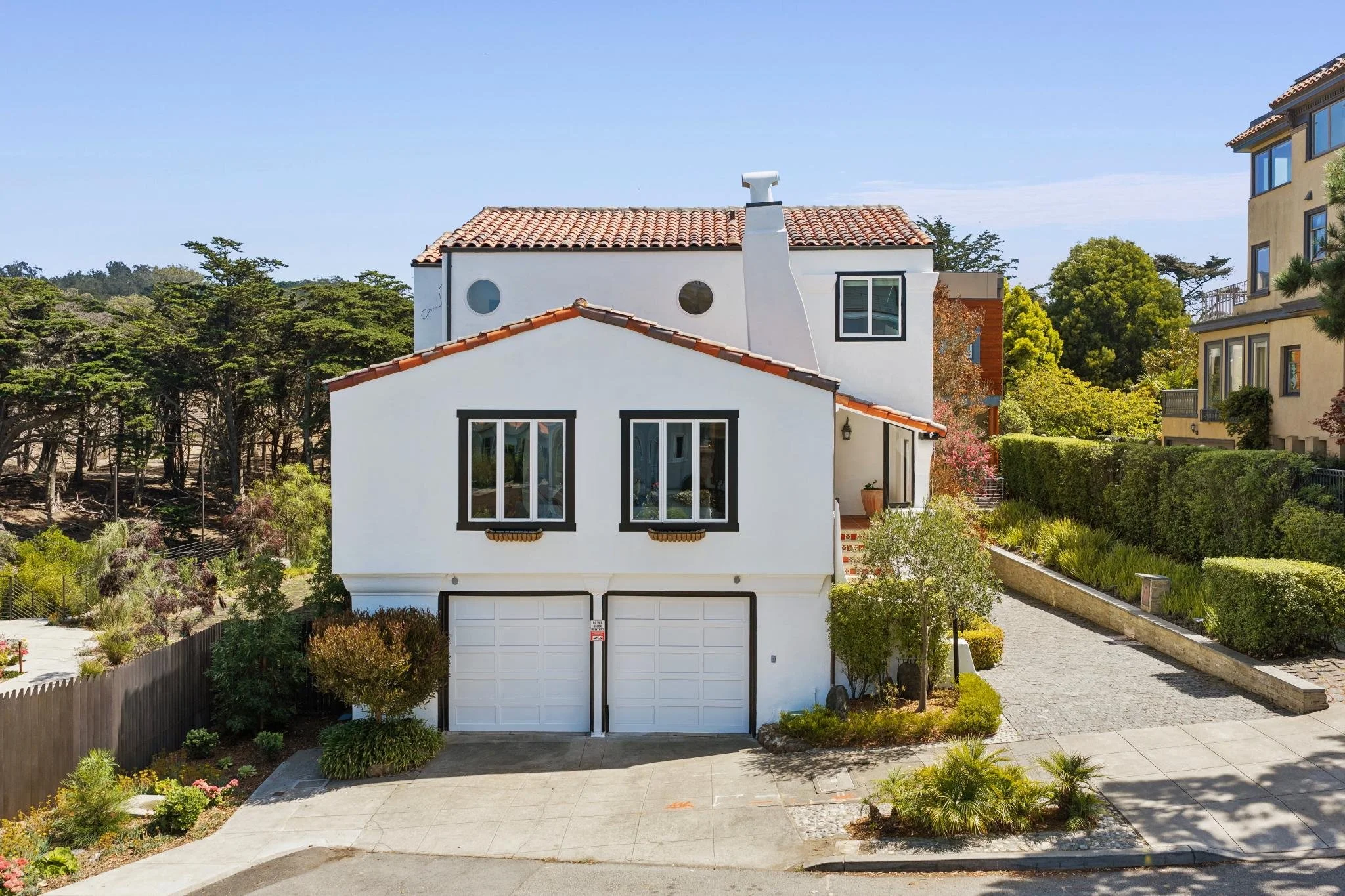 A modern white residential house with a two-car garage, black window trims, and a red-tiled roof, surrounded by landscaping and trees on a sunny day.