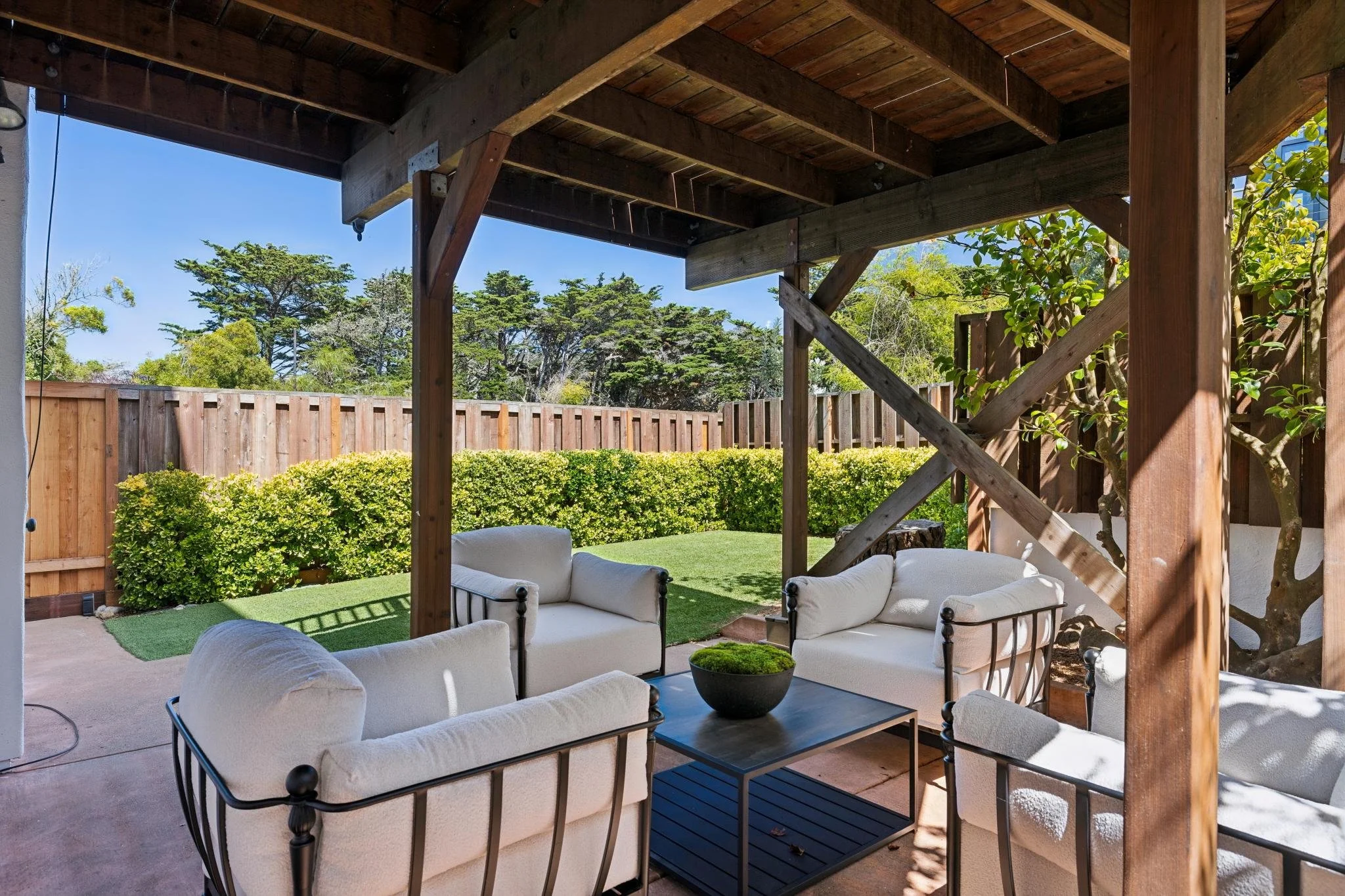 A backyard patio with outdoor seating including white cushioned chairs with black frames, a black coffee table, and a small potted plant. The patio is shaded by a wooden deck structure, with a background of green bushes, trees, and a wooden fence.