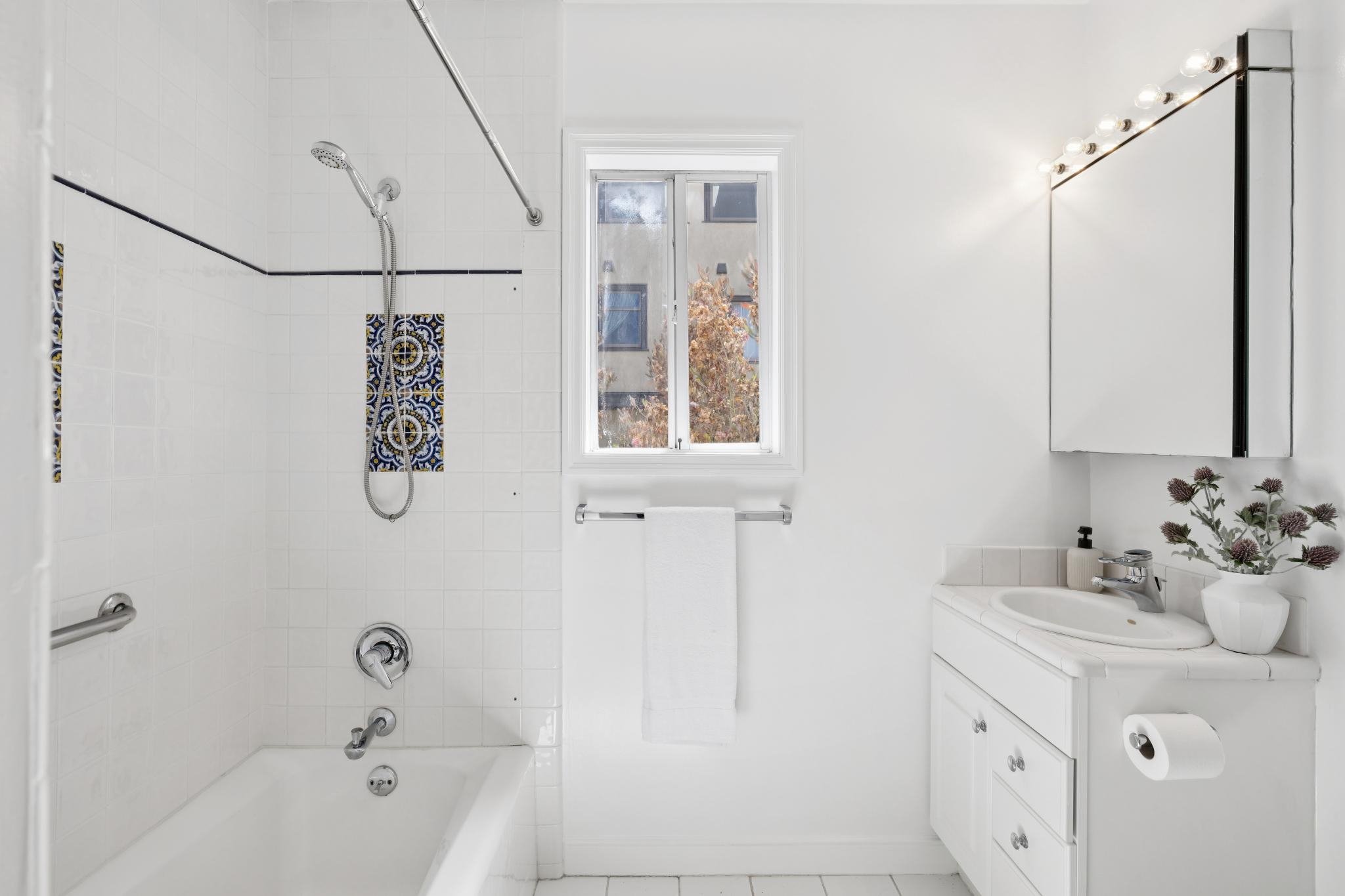A bright bathroom with a walk-in shower, a window with a view of trees outside, a white vanity with a sink, and a mirror with light bulbs. Decor includes a potted plant, soap dispenser, and hand towel.