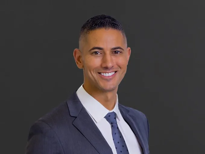 A professional man wearing a dark suit, white shirt, and polka dot tie smiling against a gray background.