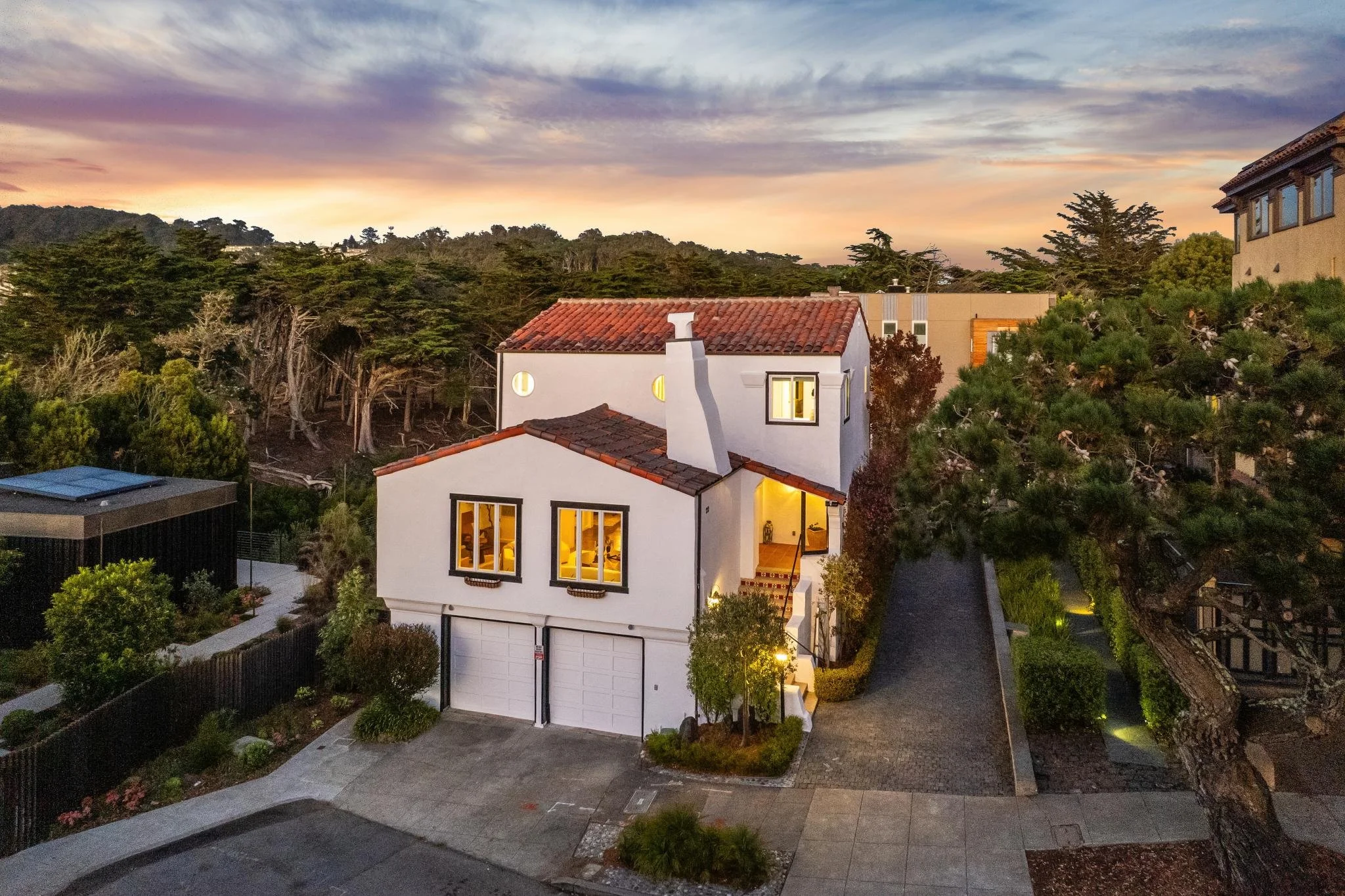 Residential neighborhood at sunset with a house that has a white exterior, a red tile roof, and large illuminated windows. There are trees and other houses nearby, with a colorful sky of pink, purple, and blue hues.