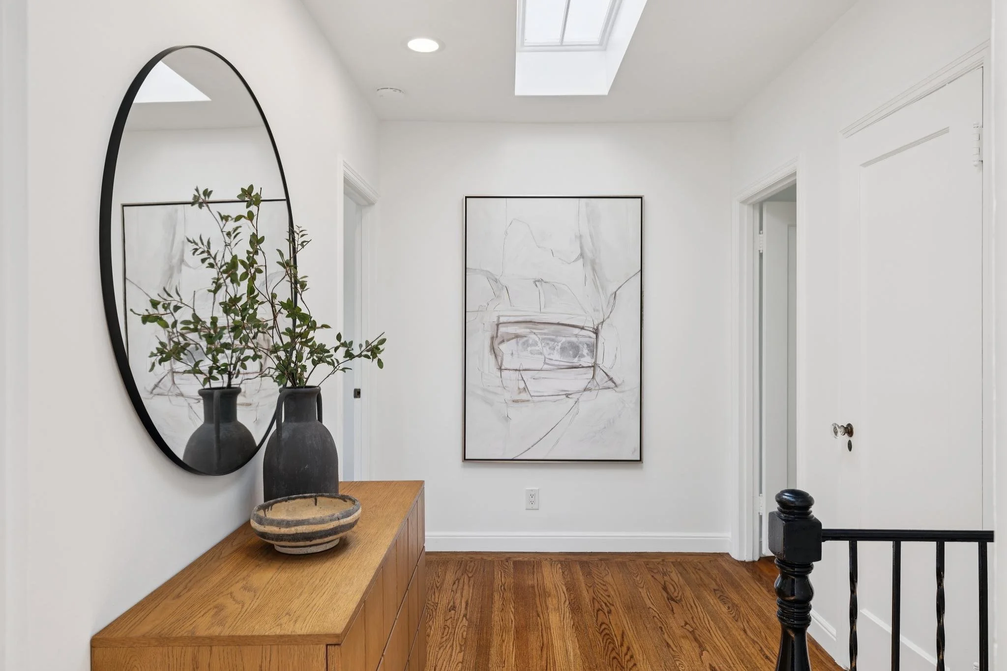 A hallway with white walls and hardwood flooring, decorated with minimalist artwork and decor. A large oval mirror hangs on the left wall reflecting some greenery in tall vases on a wooden console table. A modern abstract painting is visible on the f