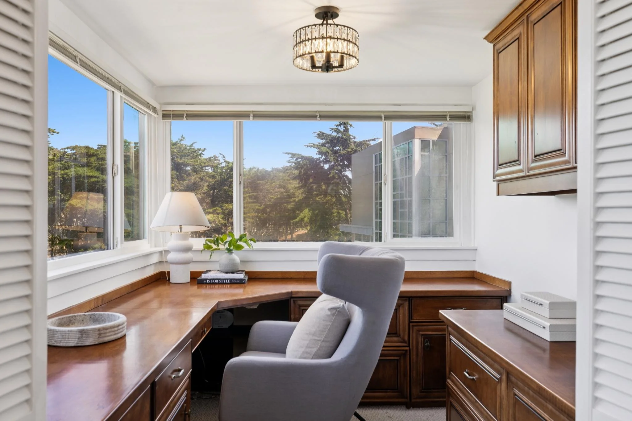 Home office with large windows showing trees outside. Wooden built-in desk with a gray chair, a white lamp, and decorative items like books and a plant. Ceiling light fixture.
