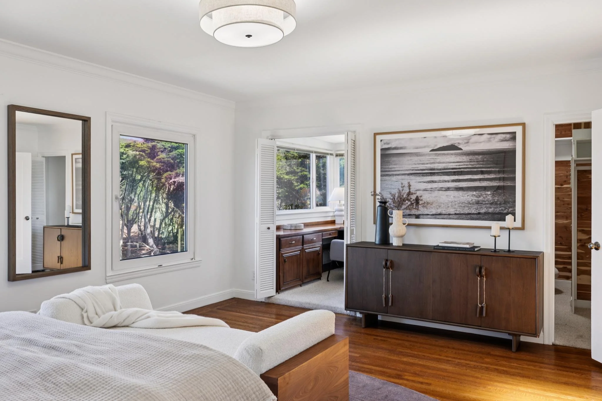 A living room with white walls, hardwood floors, a white couch, a large black and white ocean photo on the wall, a wooden sideboard with decorative vases and books, and a window with trees outside.