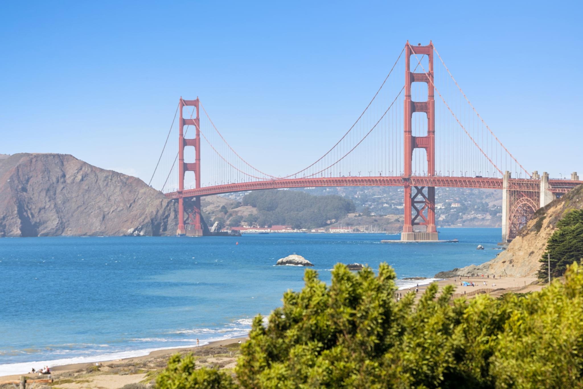 View of the Golden Gate Bridge spanning the water, with hills and a clear blue sky in the background. Green foliage and a beach are in the foreground.