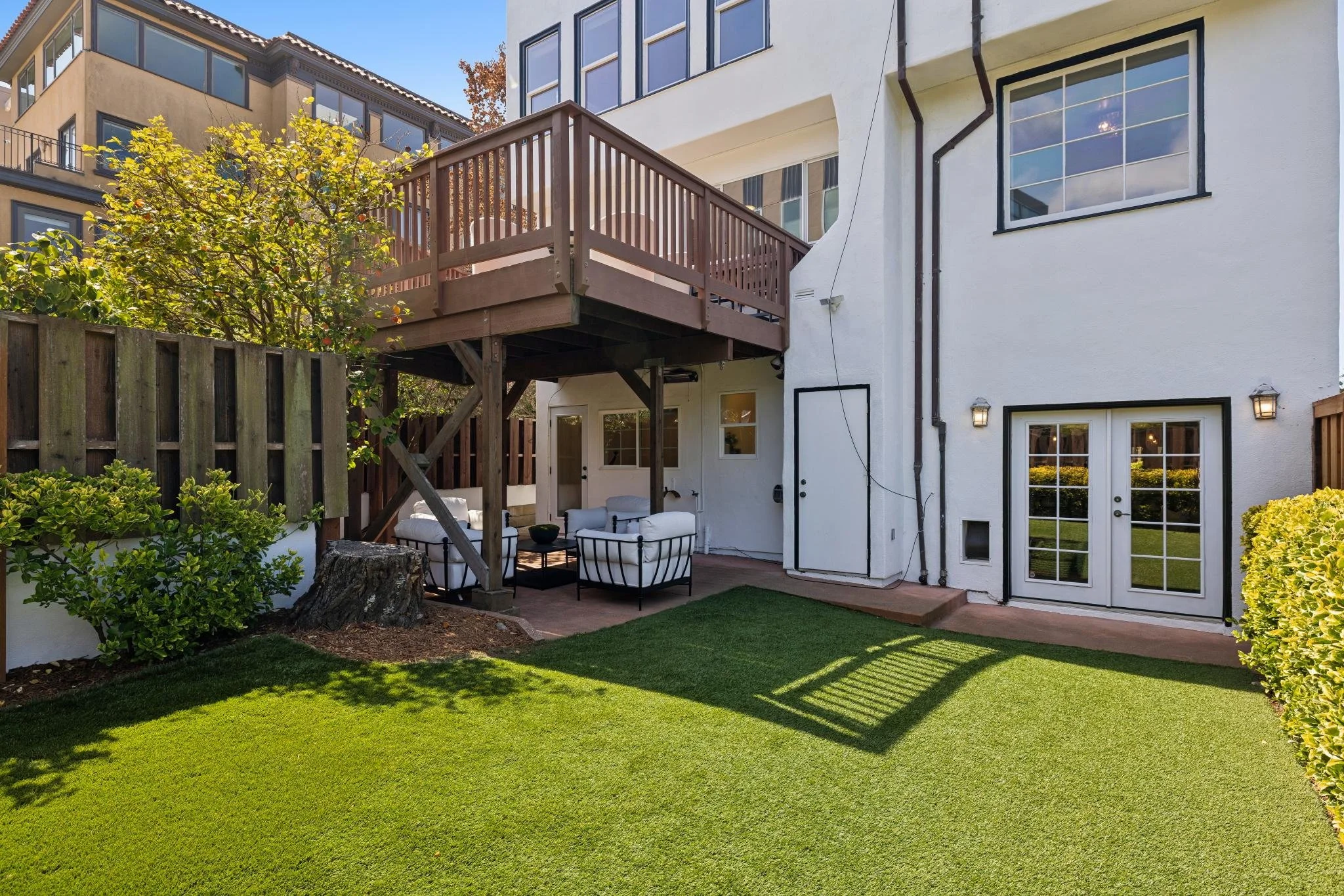 Backyard patio with white outdoor furniture, a wooden deck, lush green lawn, shrubbery, and a white house with large windows.