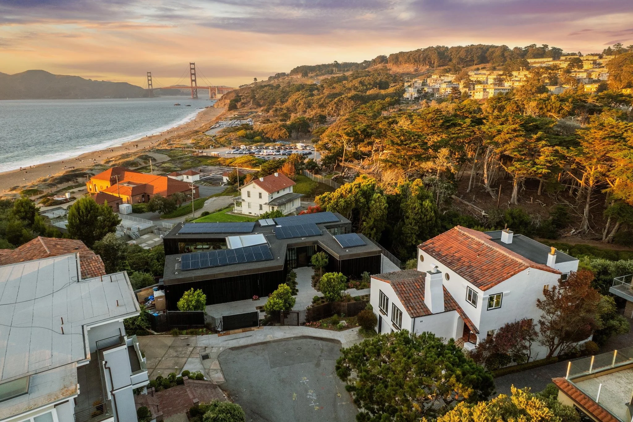 A view of a coastal neighborhood at sunset featuring houses, trees, a beach, and a bridge in the background.