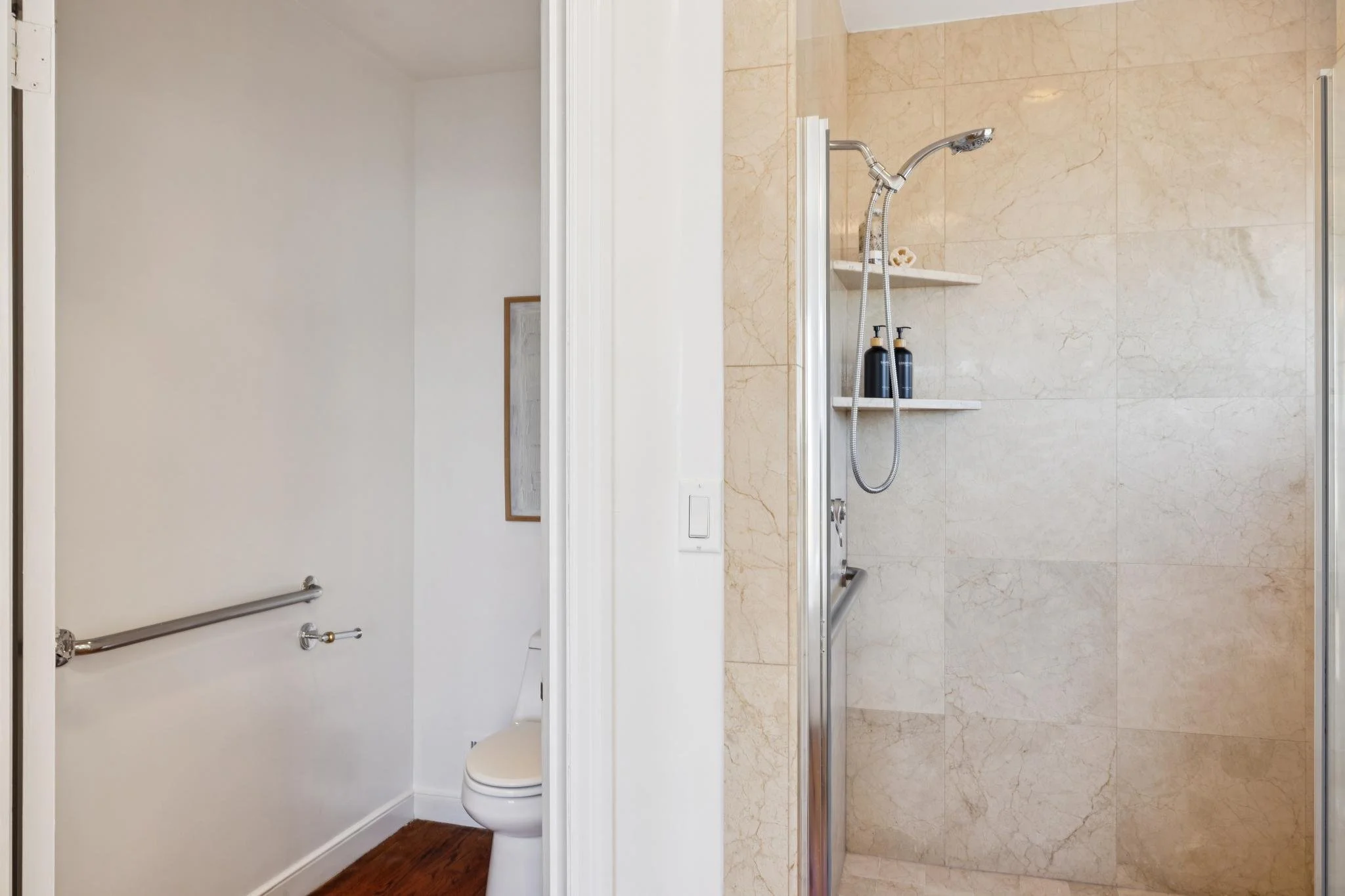 Bathroom with a walk-in shower with beige marble tiles, a chrome shower head, and three black bottles on a built-in shelf. Next to the shower is a white wall with a toilet and a handrail.