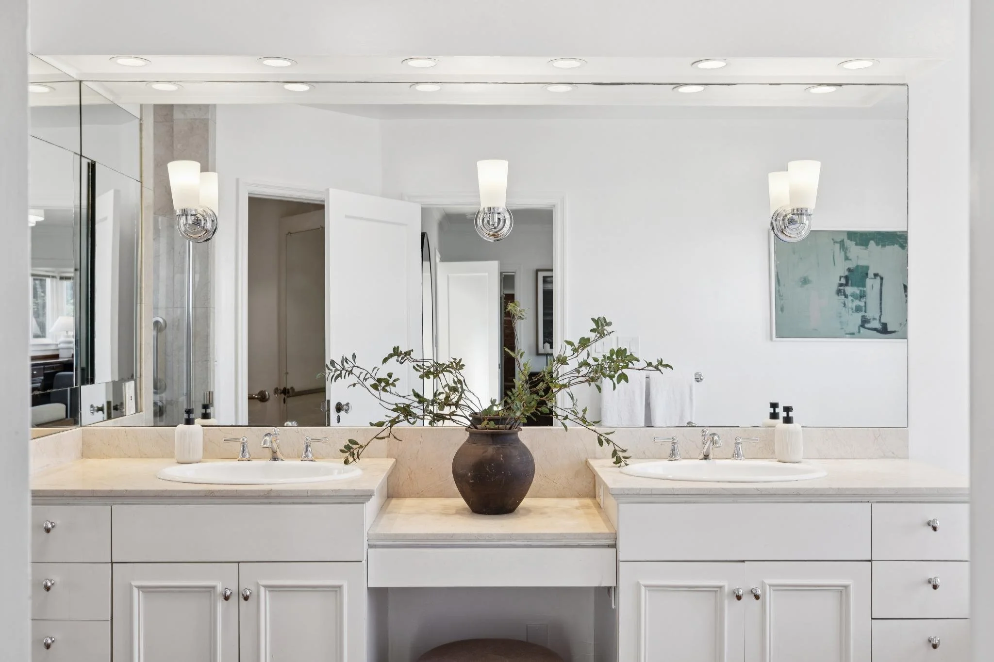 A modern bathroom vanity with a large mirror, two sinks, and a potted plant in the center. The wall has three light fixtures, and there are paintings and an open door in the background.