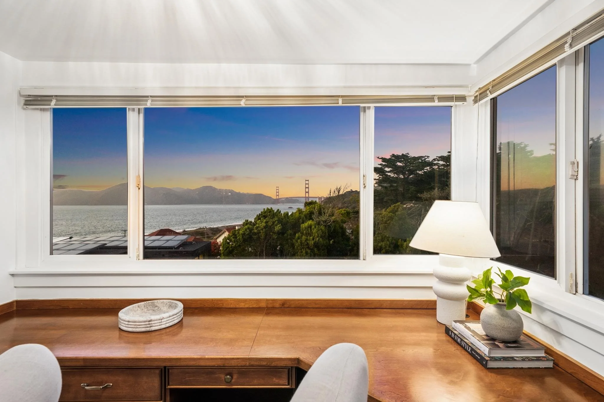 Interior view of a room with a large window showing a sunset view of a body of water, mountains, and a bridge, decorated with a white table lamp, potted plant, and books.