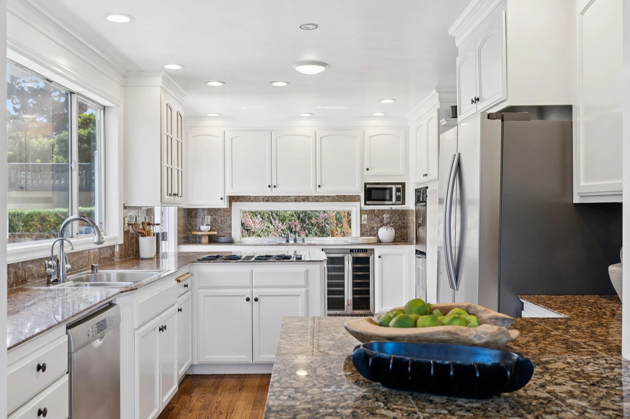 Modern kitchen with white cabinets, granite countertops, stainless steel appliances, and hardwood flooring. A window above the sink overlooks greenery. There are recessed ceiling lights and a bowl of green apples on the counter.