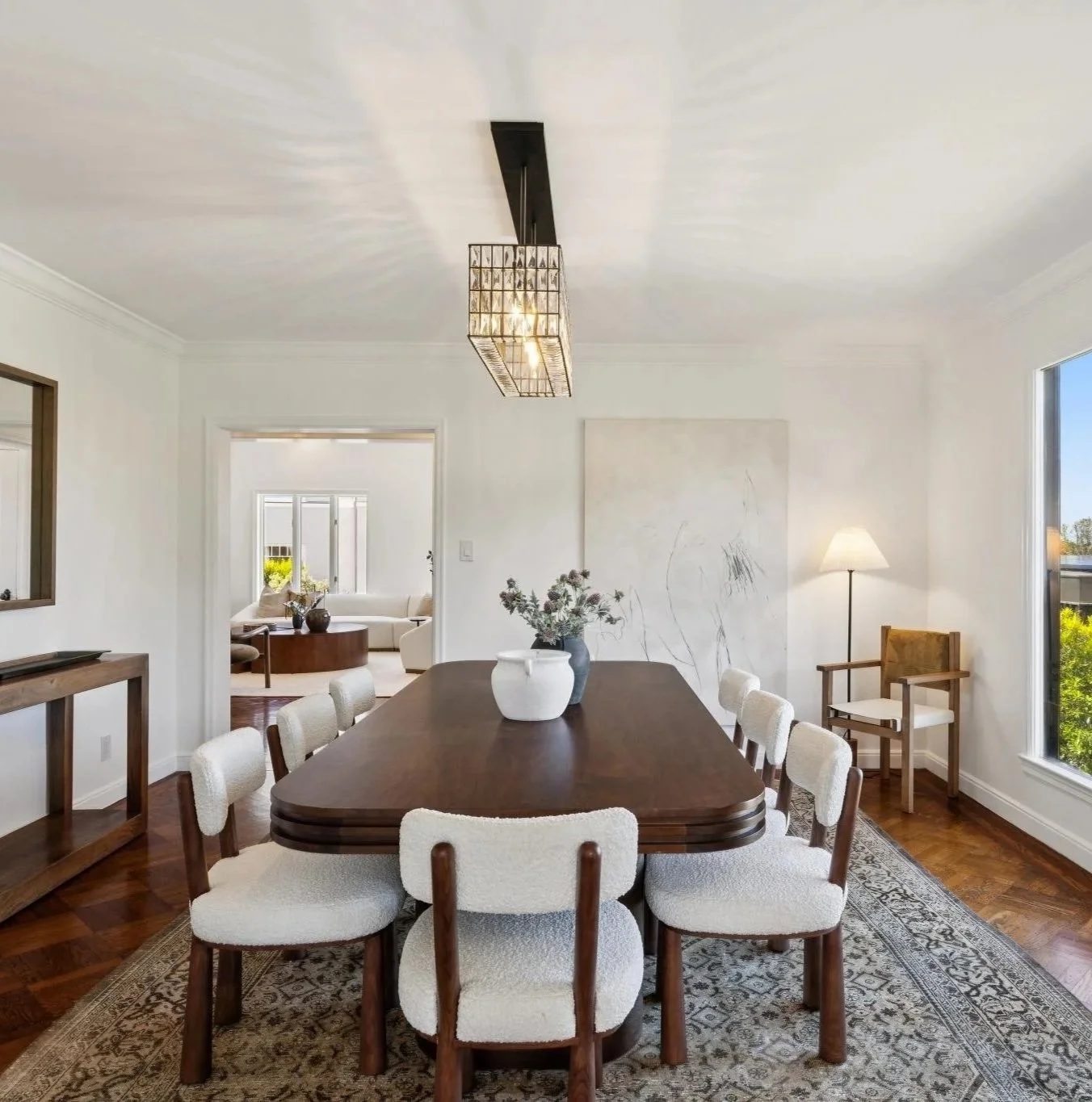 A modern dining room with a dark wooden table, white upholstered chairs, a patterned area rug, and a large window showing greenery outside. A white vase with flowers sits on the table, and the room is lit by a rectangular chandelier.