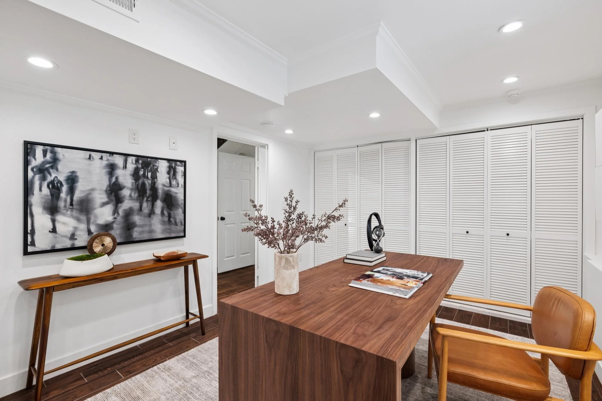 Modern dining room with a wooden table, orange chair, decorative branches in a vase, and wall art of blurred black-and-white people, white walls, and white louver closet doors.