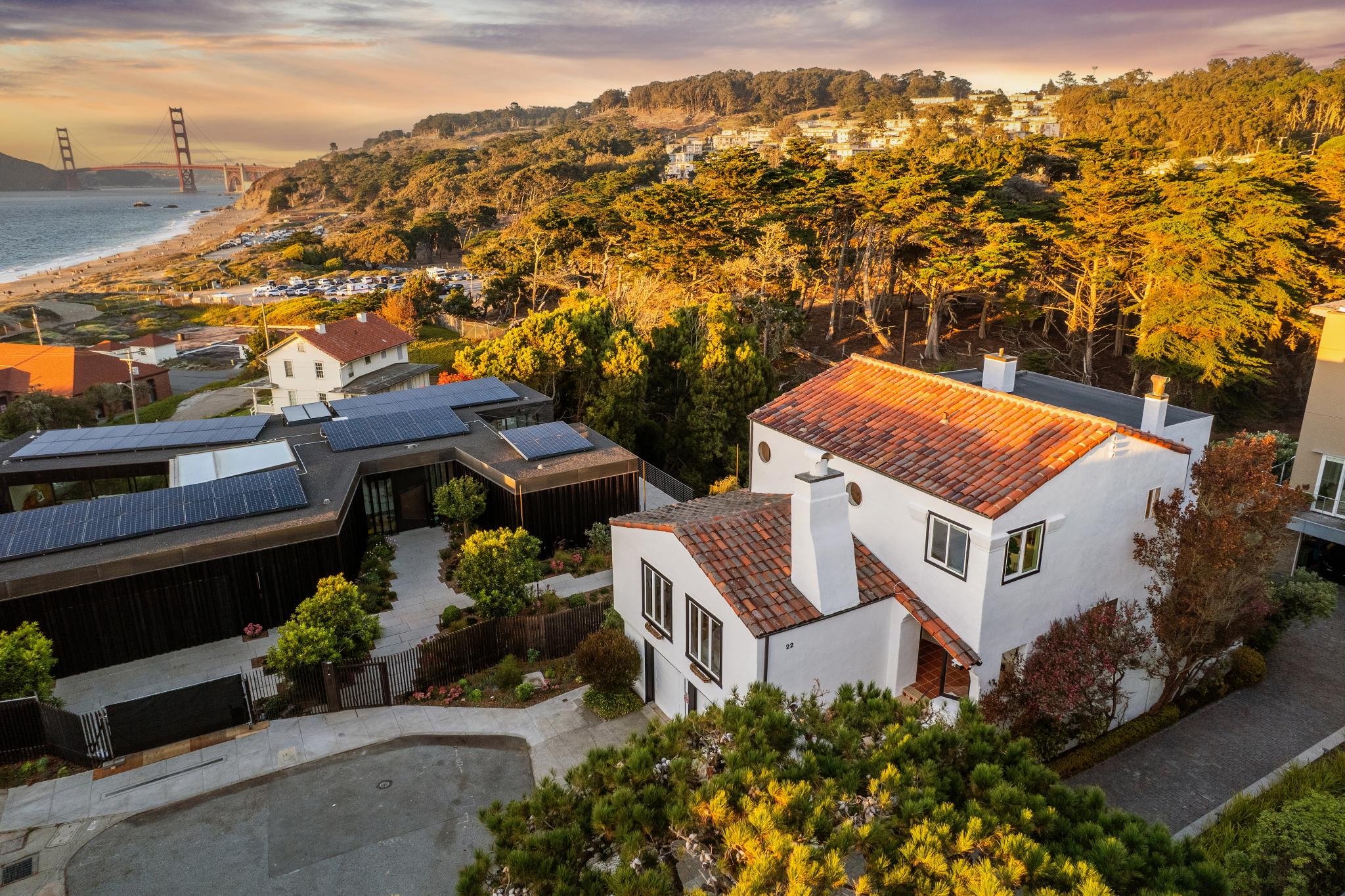 Overhead view of a coastal neighborhood with houses, trees, and a parking lot, with the Golden Gate Bridge in the background during sunset.