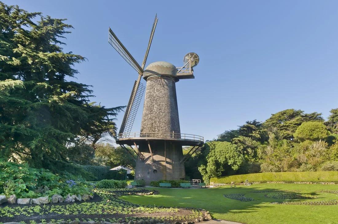 A traditional windmill with four blades standing in a lush green garden under a clear blue sky.