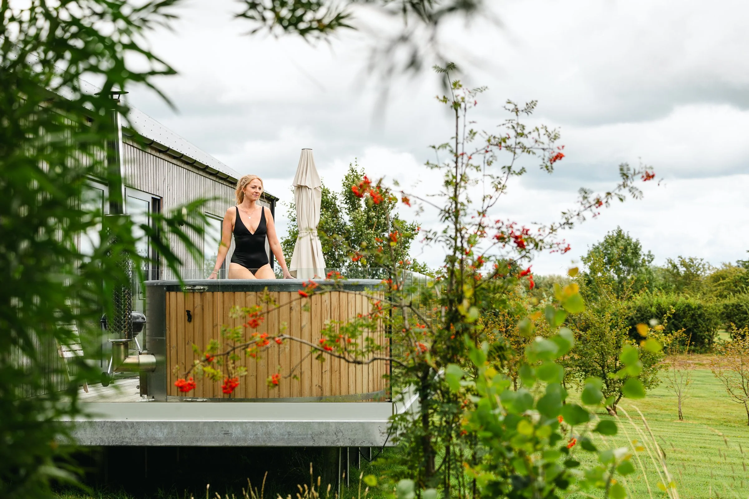 A woman in a black swimsuit is sitting on the edge of a wooden hot tub outdoors surrounded by greenery and flowering plants, with a cloudy sky overhead.