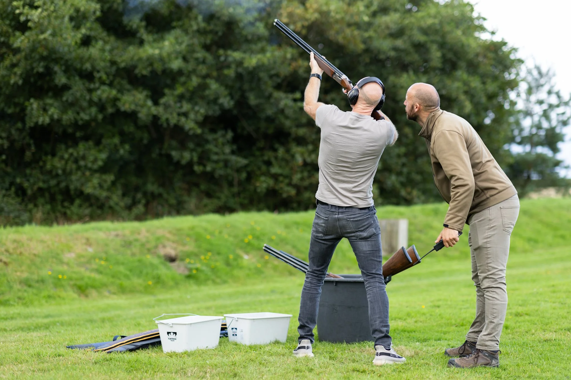 Two men at a shooting range with shotguns, one aiming and the other observing, on a grassy outdoor area with trees in the background.
