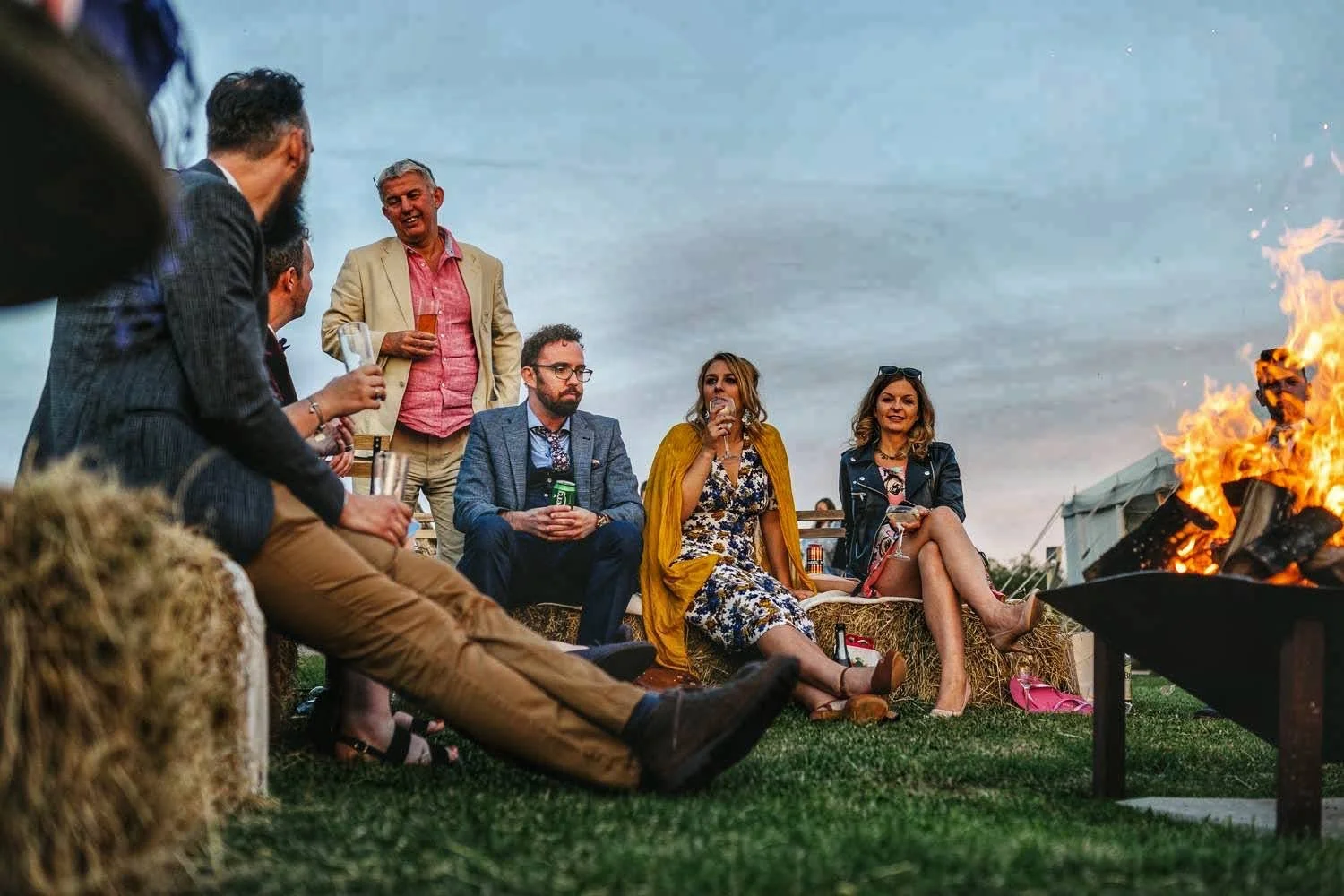 A group of people sitting around a bonfire outdoors during sunset, some holding drinks, socializing and enjoying the moment.