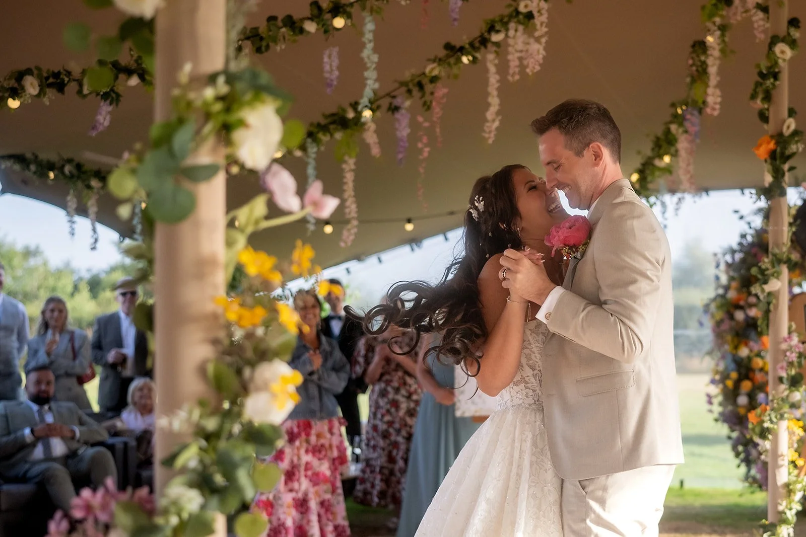 A bride and groom dancing at their wedding under a floral decorated canopy with guests watching and smiling in the background.