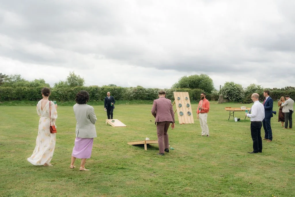 Group of people playing cornhole outdoors on a cloudy day with green grass and bushes in the background.