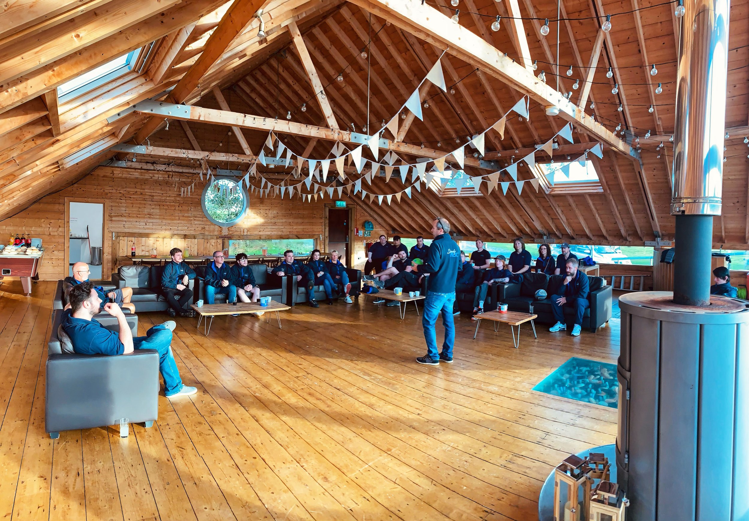 People gathered in a wooden event space with bunting decorations and string lights, seated on couches and chairs, listening to a speaker standing in front of them.