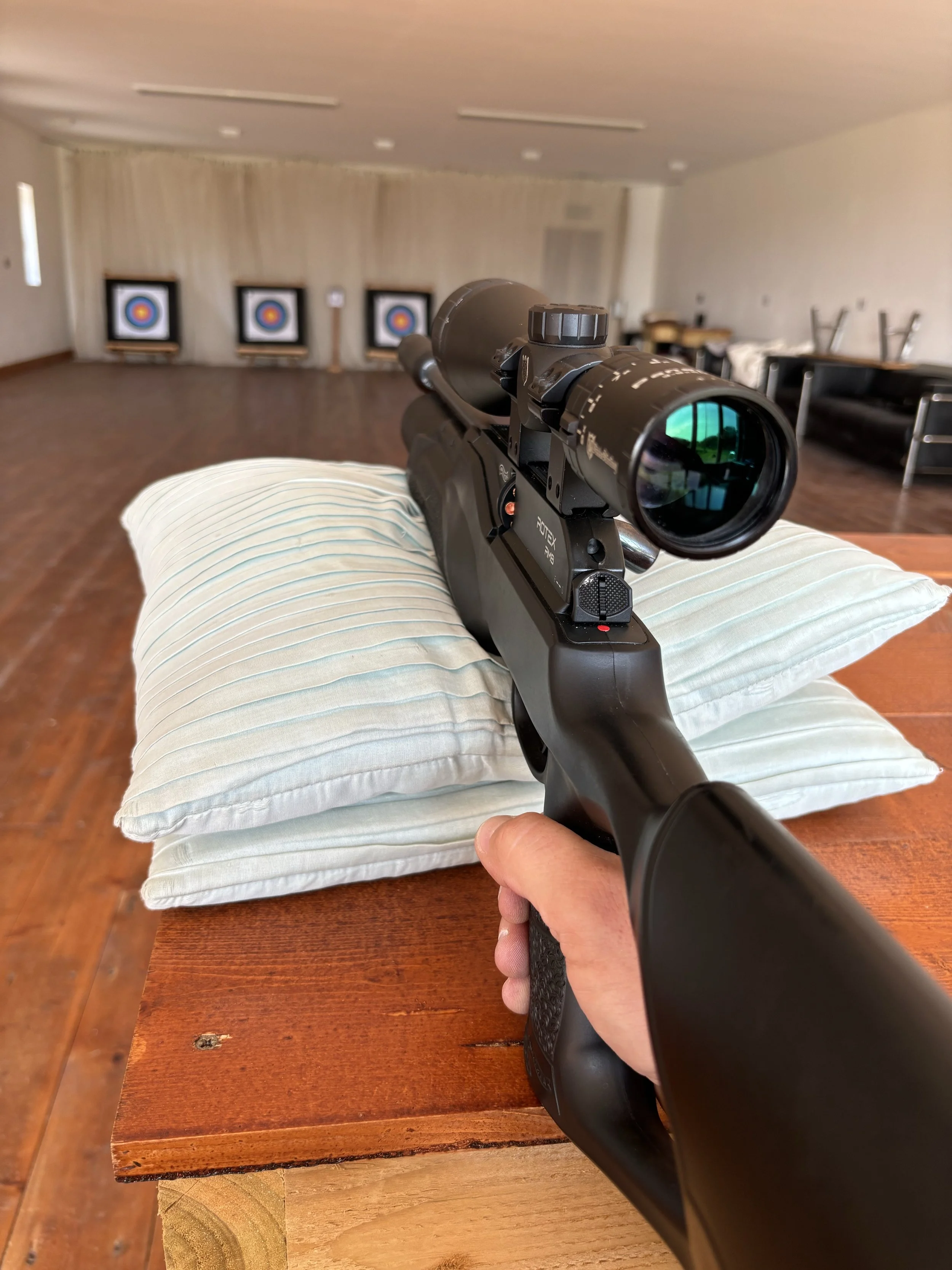 A person holding a black rifle with a scope, pointing it towards a row of three archery targets in a room with wooden floors and beige curtains.