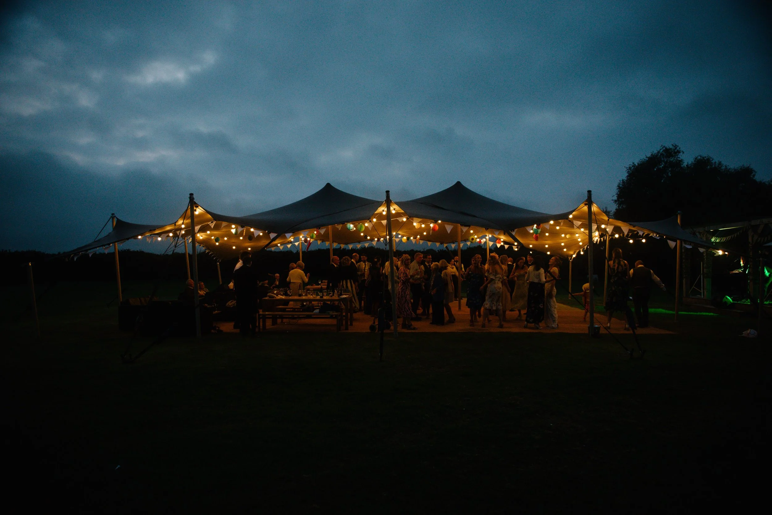 People gathered under a tent decorated with string lights and bunting at dusk outdoor event.