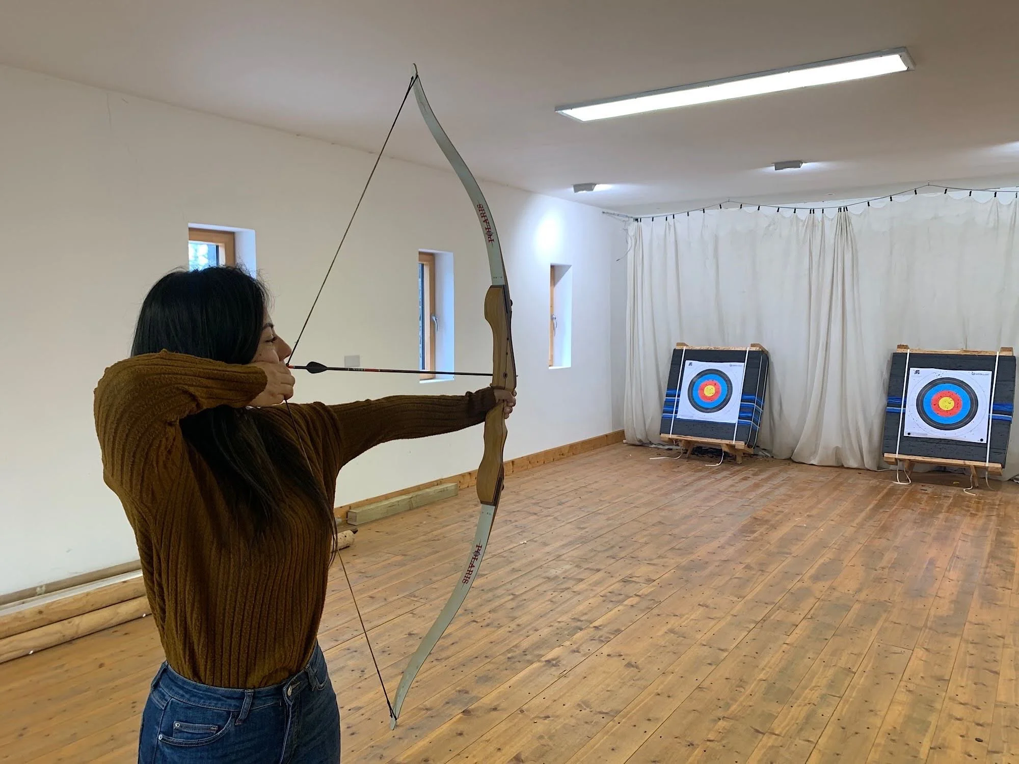 A woman in a brown sweater and blue jeans is aiming an arrow from a bow in an indoor archery range with archery targets in the background.