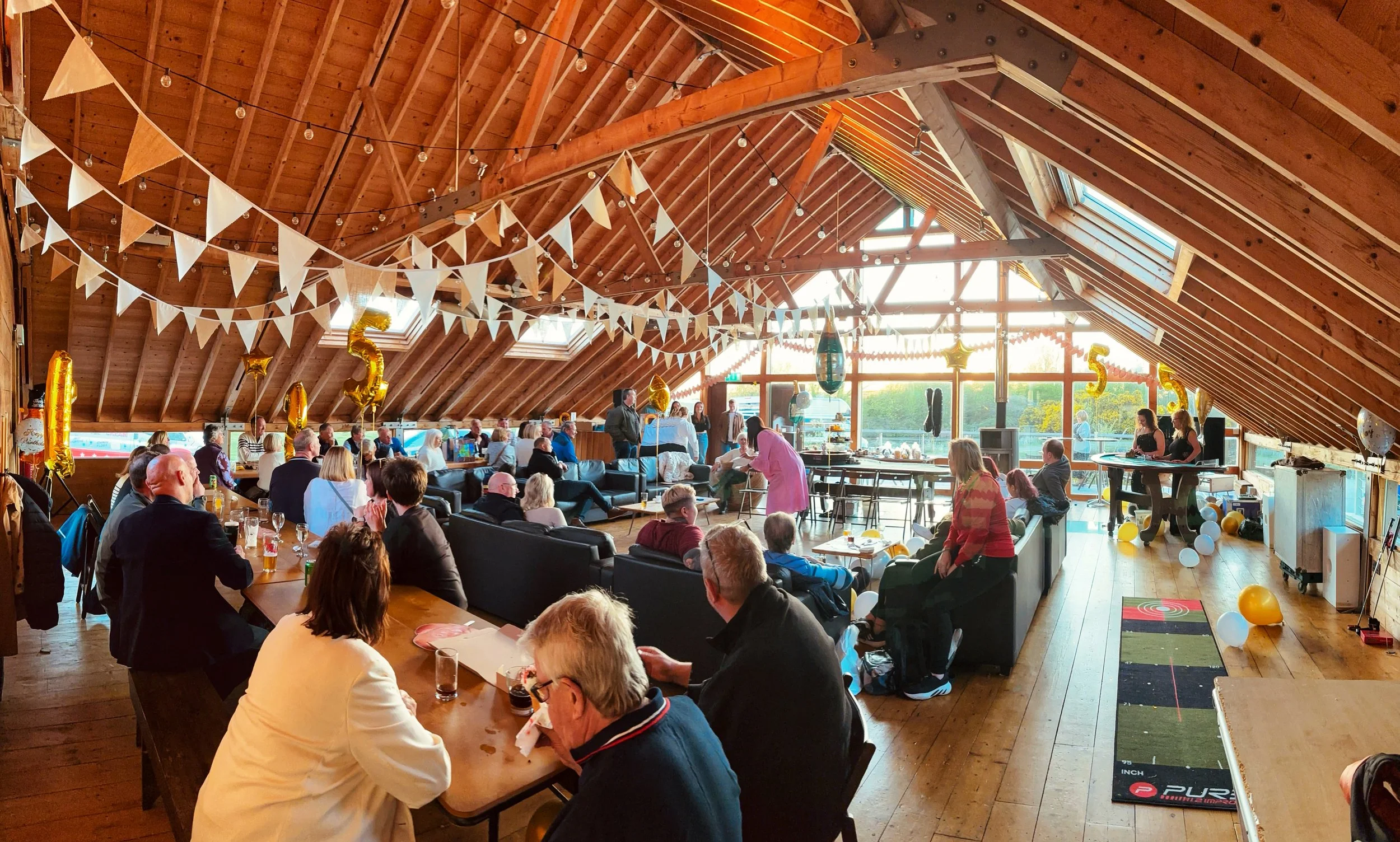 A gathering of people inside a decorated wooden venue with string lights, white and gold balloons, and bunting banners. There are multiple tables, some people sitting and some standing, celebrating a special occasion, possibly a birthday, with a large window letting in natural light.