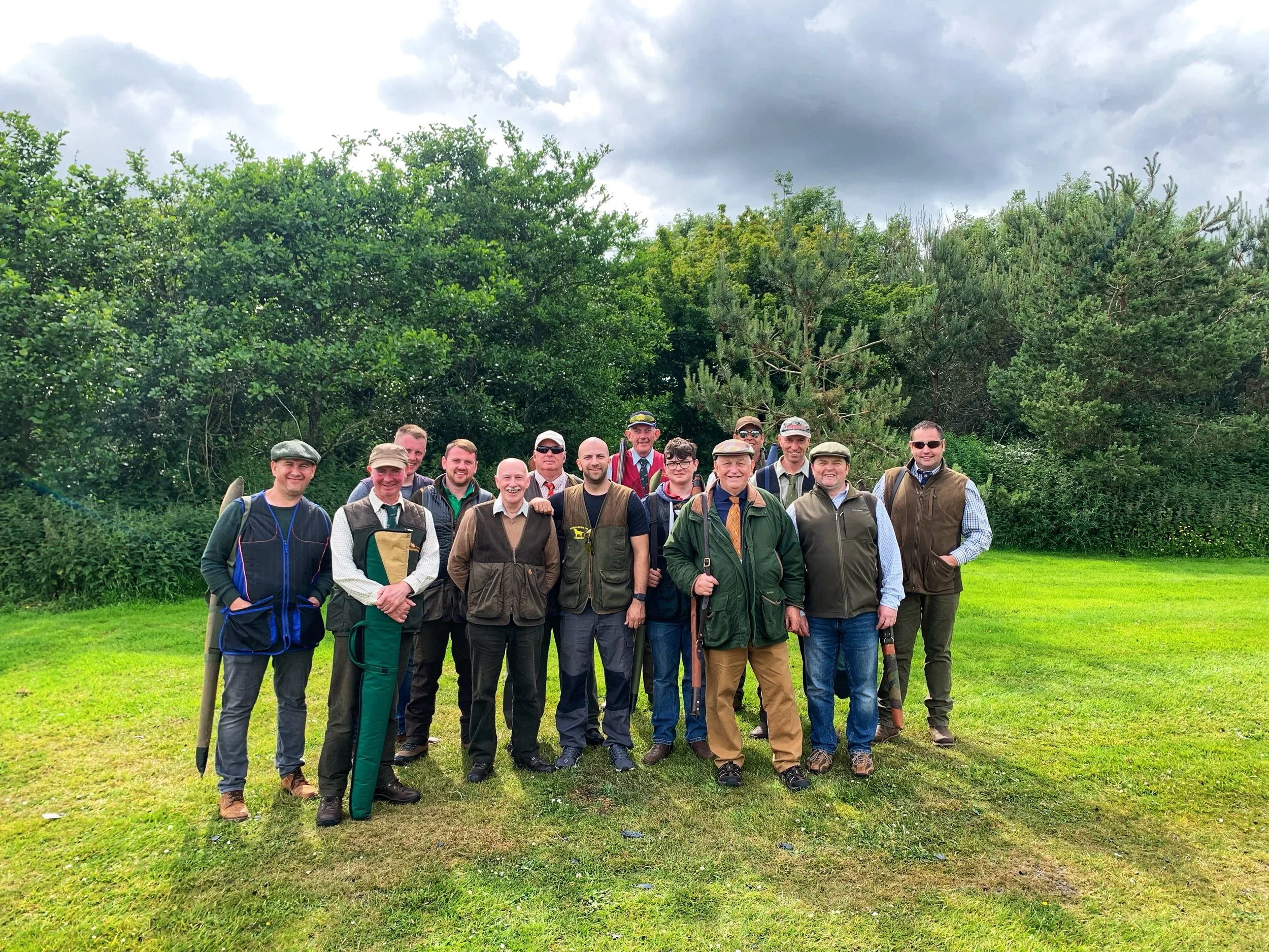 A group of around twenty men and women standing together outdoors on a grassy area, with green trees and bushes behind them. The sky is cloudy. The individuals are dressed in outdoor and hiking clothing, some with hats and backpacks, and appear to be posing for a photo.