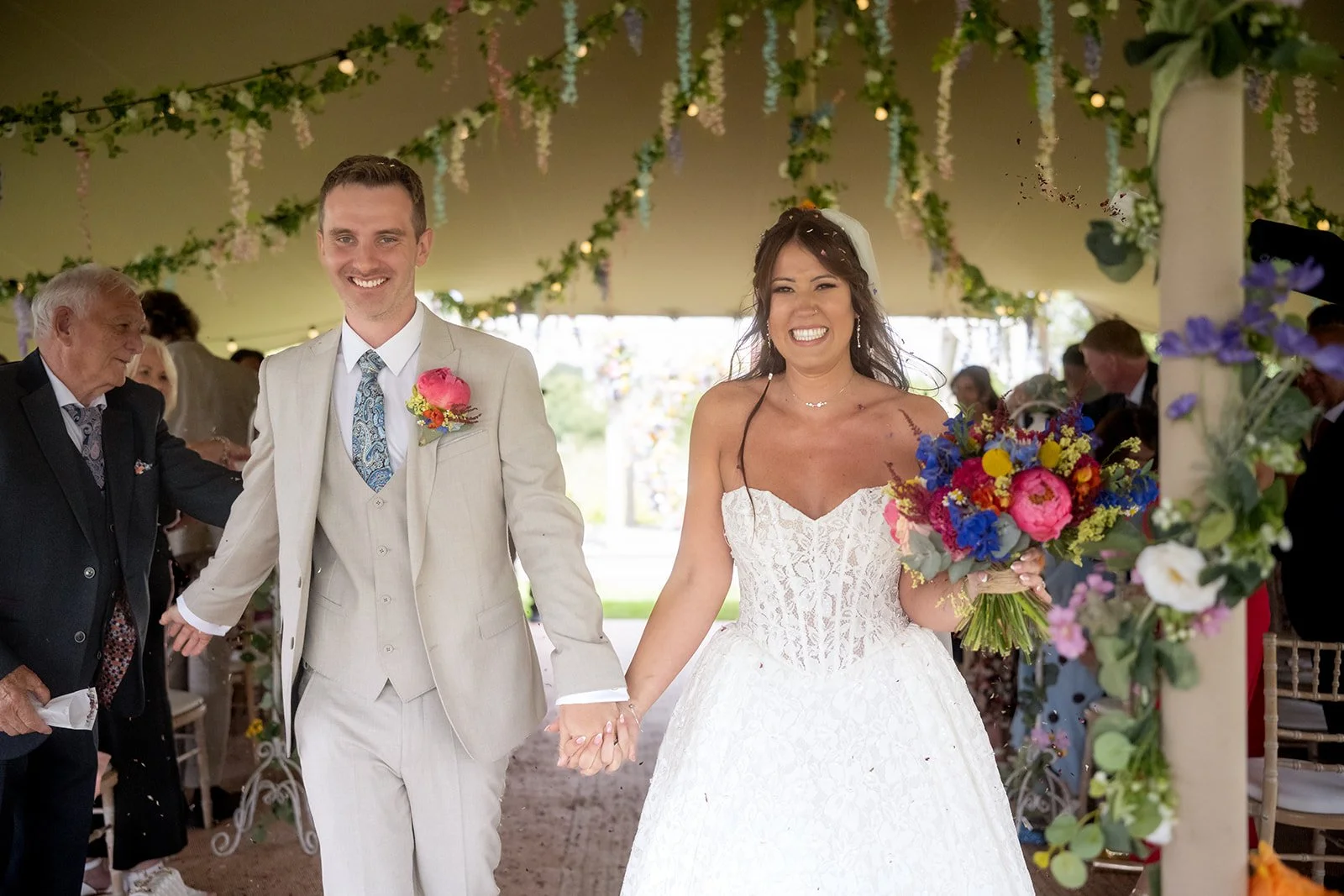A newlywed couple walking hand in hand at their wedding reception under a decorated canopy, smiling happily. The bride holds a vibrant bouquet of pink, yellow, and blue flowers. The groom is dressed in a light-colored suit with a floral tie and bouto