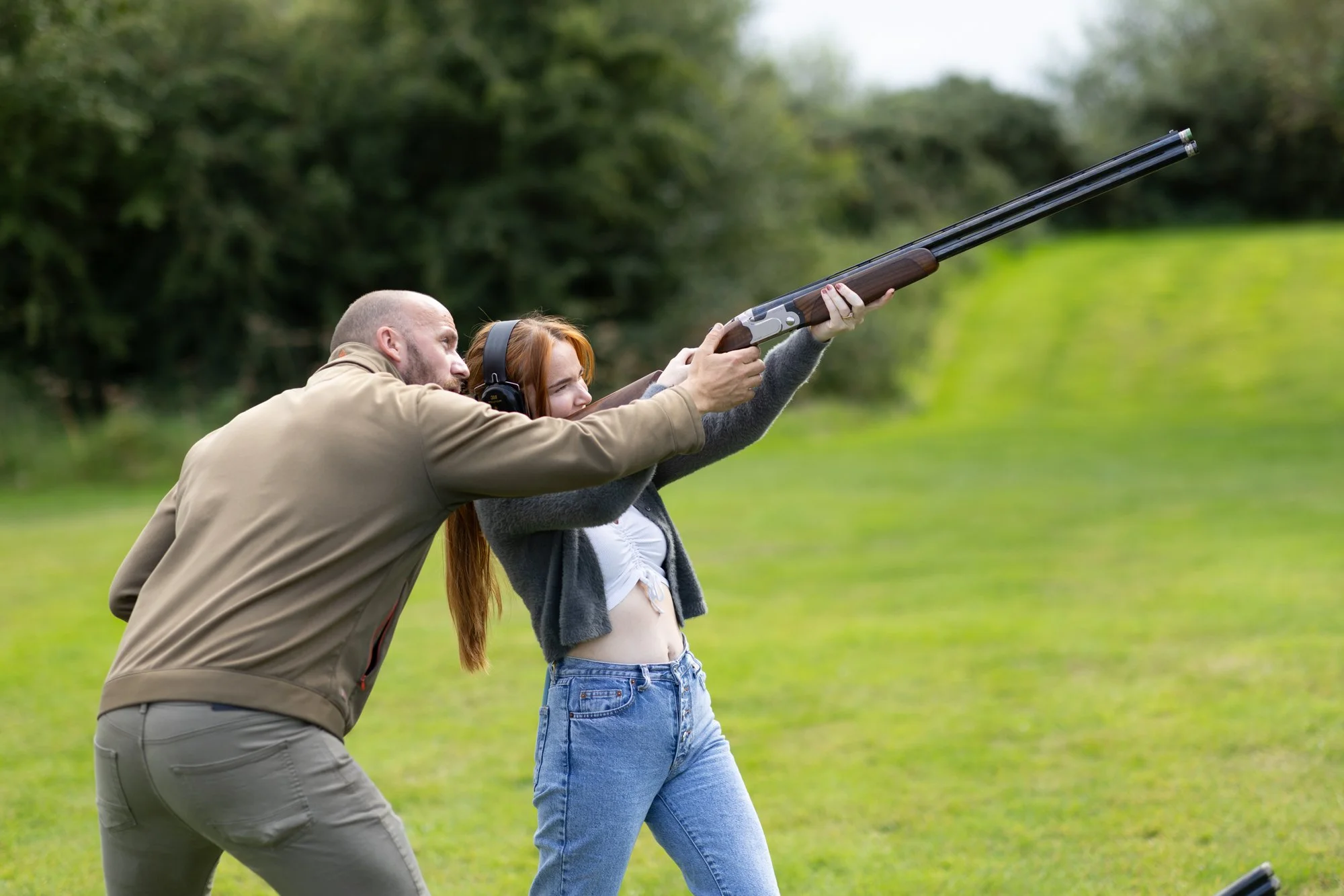 A man and a young woman practicing shooting with a shotgun outdoors on a grassy field, with trees in the background.
