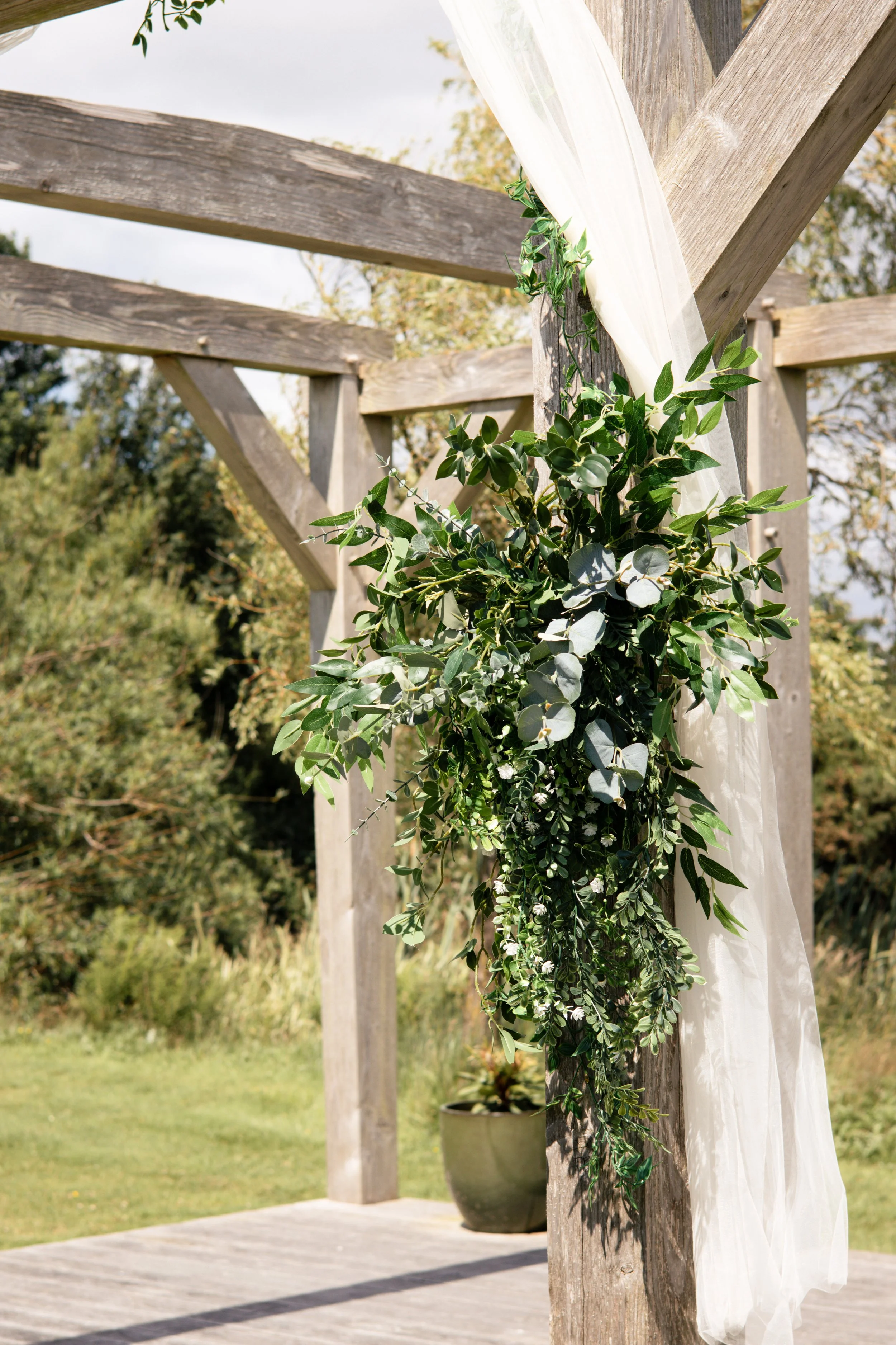 Decorative outdoor setup with a white fabric hanging from wooden posts adorned with green leafy garlands, on a wooden platform with a grassy background.