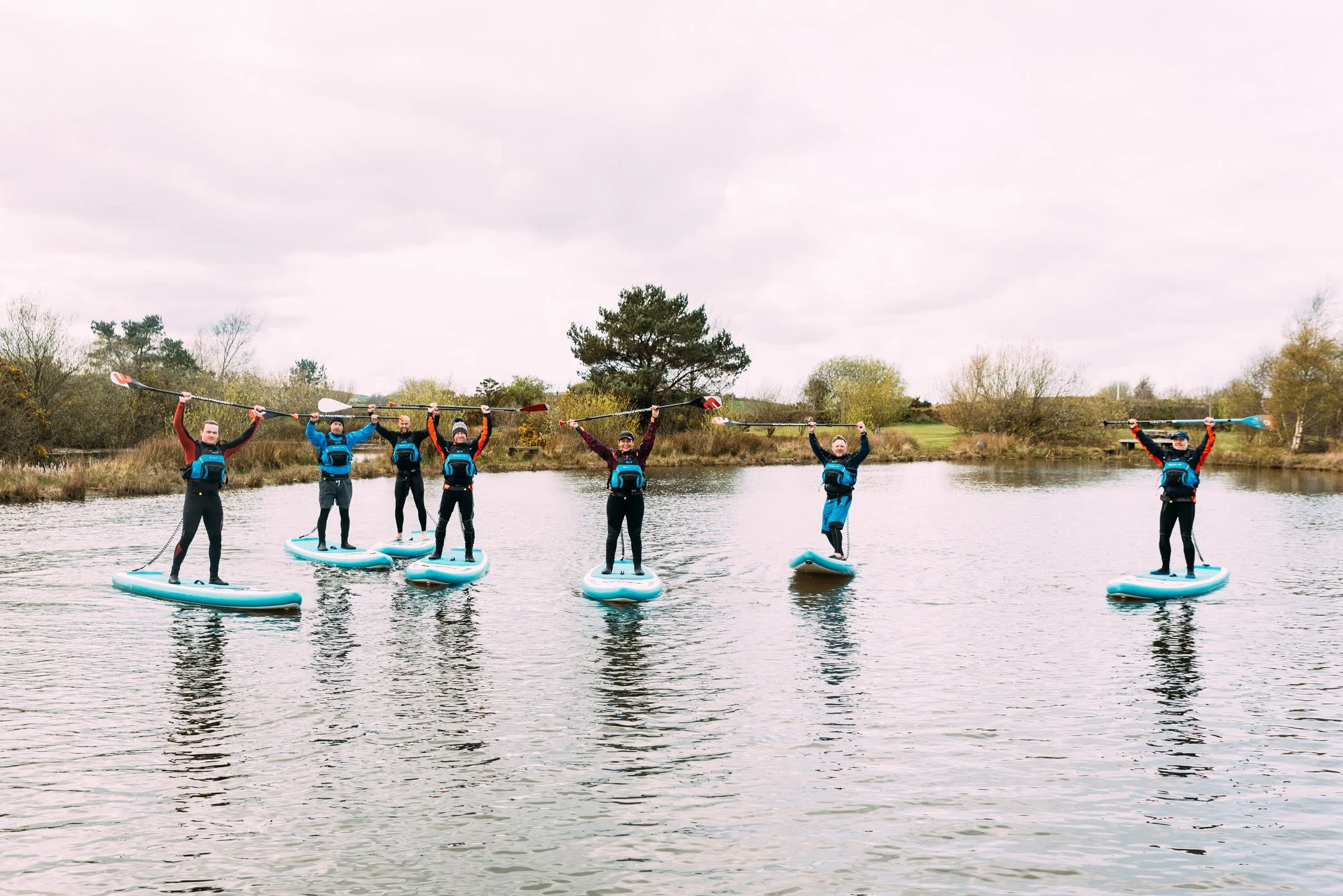 Six people standing on paddleboards on a lake, holding paddles above their heads, with trees in the background under a cloudy sky.