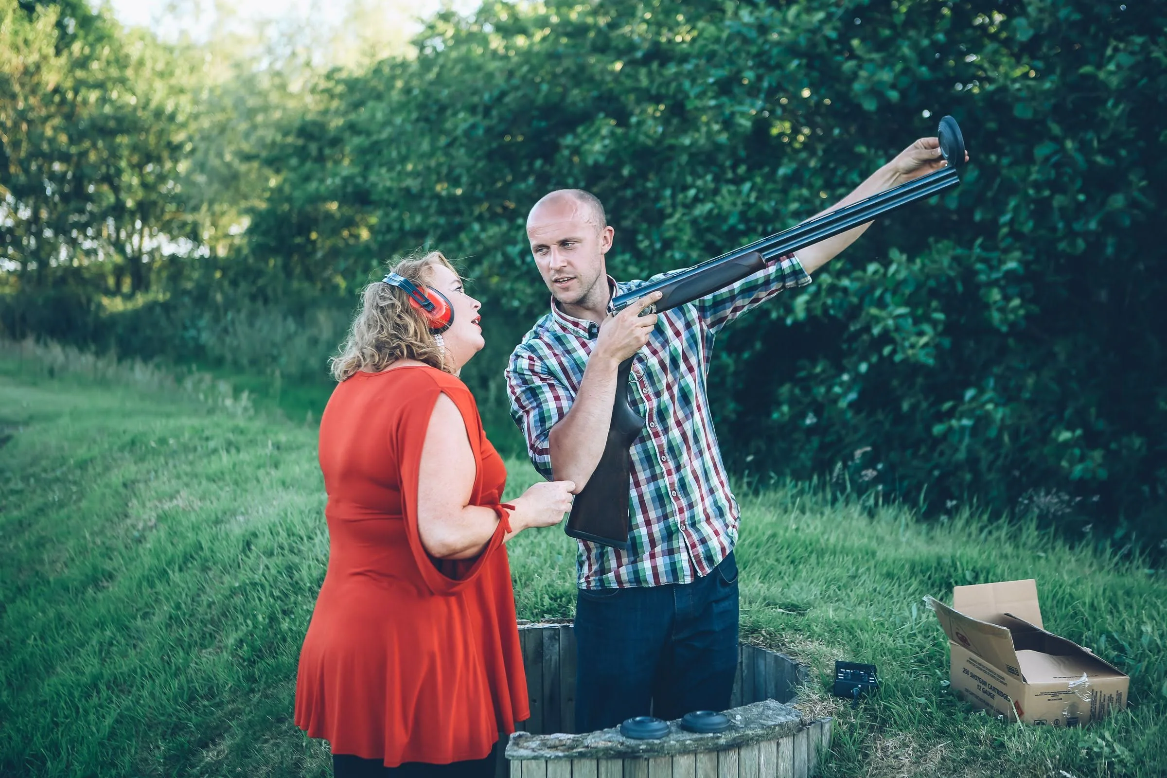 A man and woman outdoors with trees in the background, the man is holding a shotgun and the woman is wearing ear protection, both are engaged in conversation.
