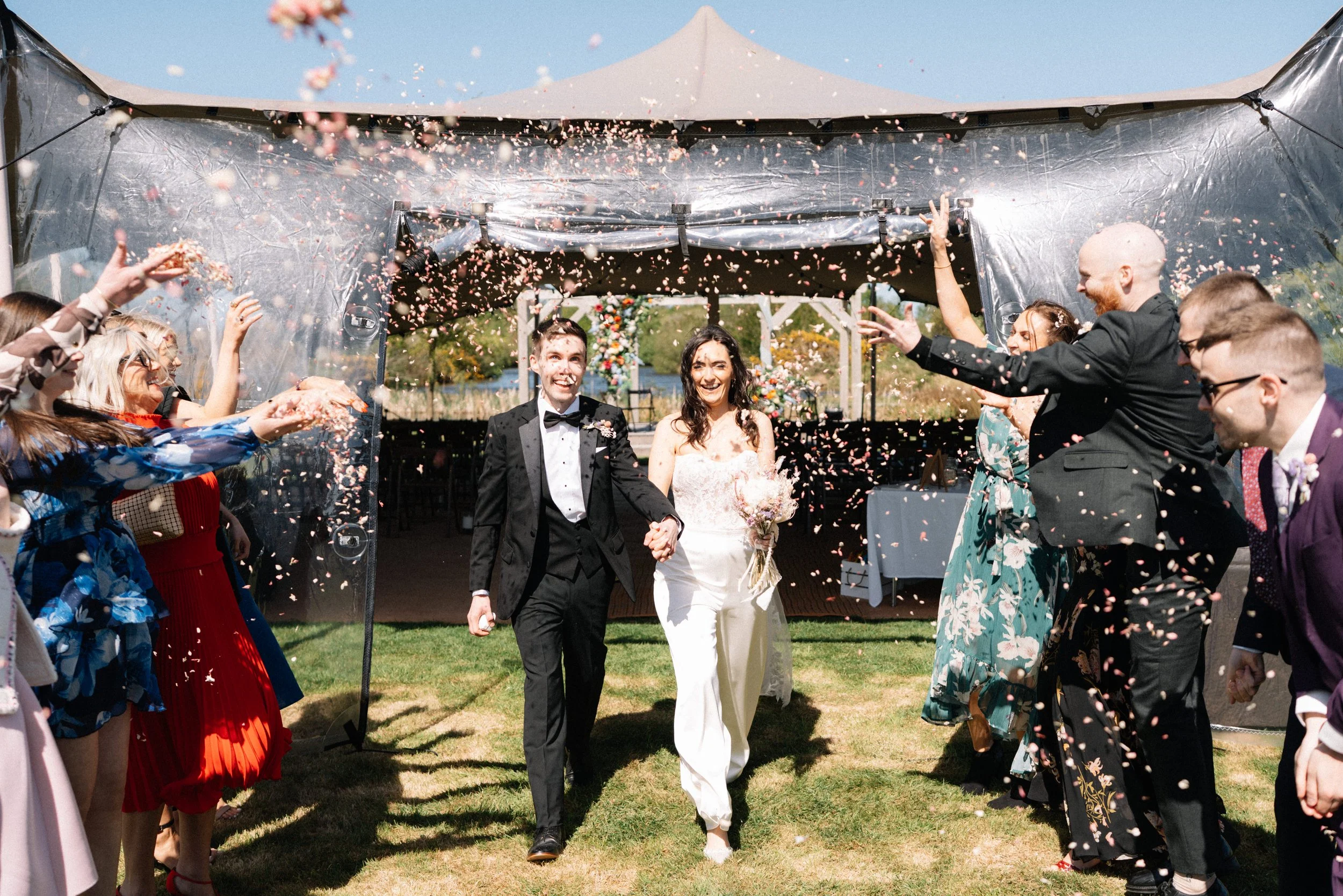A newlywed couple walking hand in hand through friends and family celebrating at an outdoor wedding reception, with confetti falling around them.