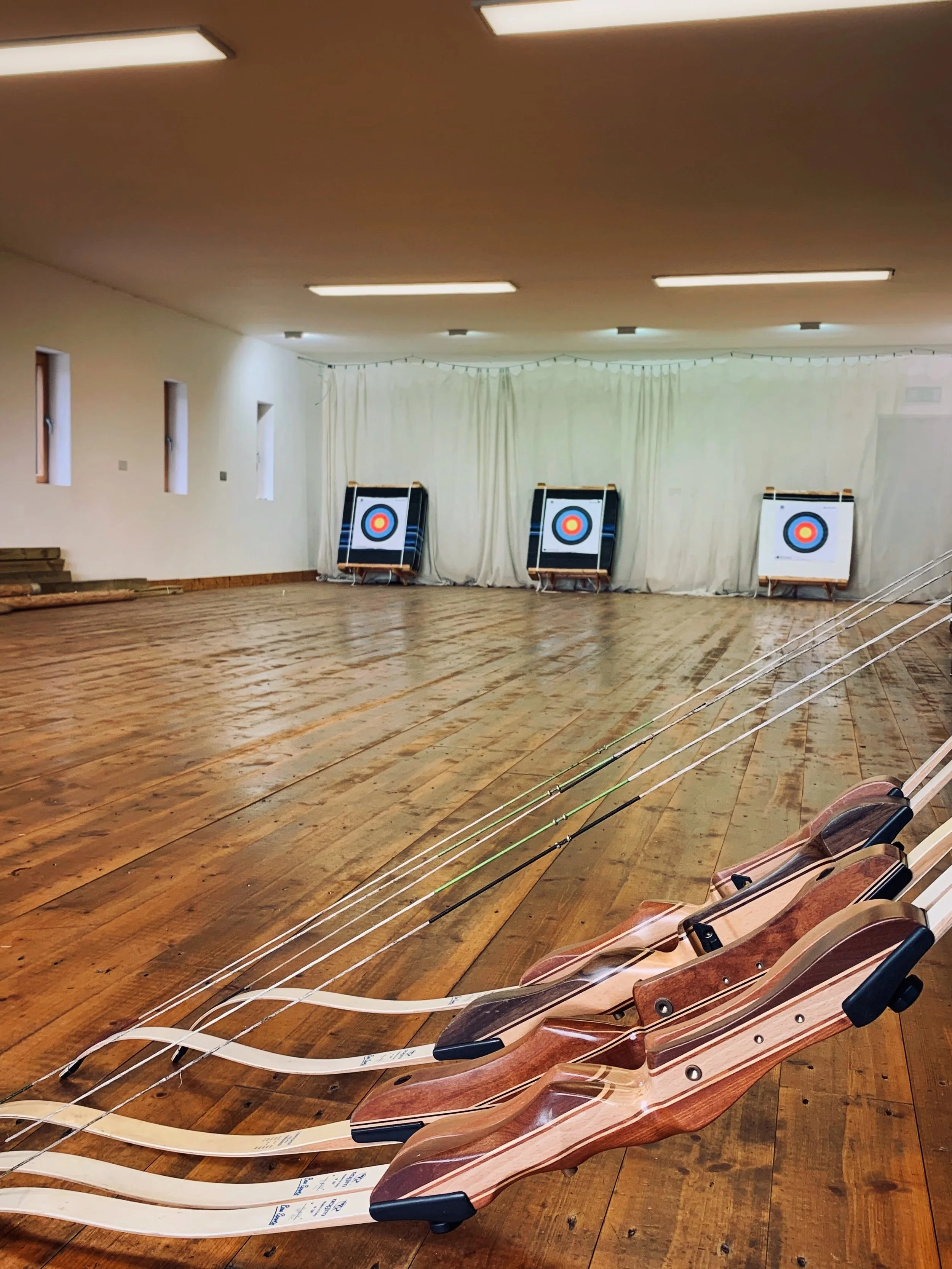 Indoor archery range with three archery targets set up at the back wall and several wooden bows with white arrow shafts lined up on the floor in the foreground.