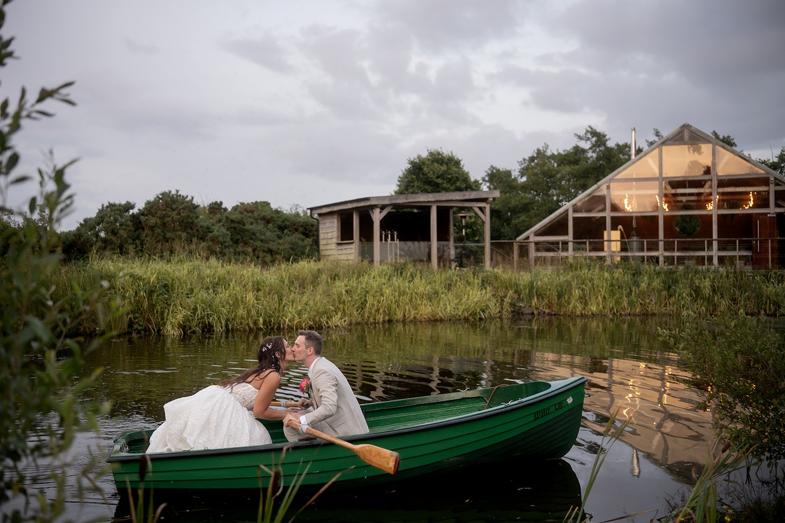 A bride and groom kissing in a green rowboat on a calm lake during sunset, with a wooden cabin and lush trees in the background.