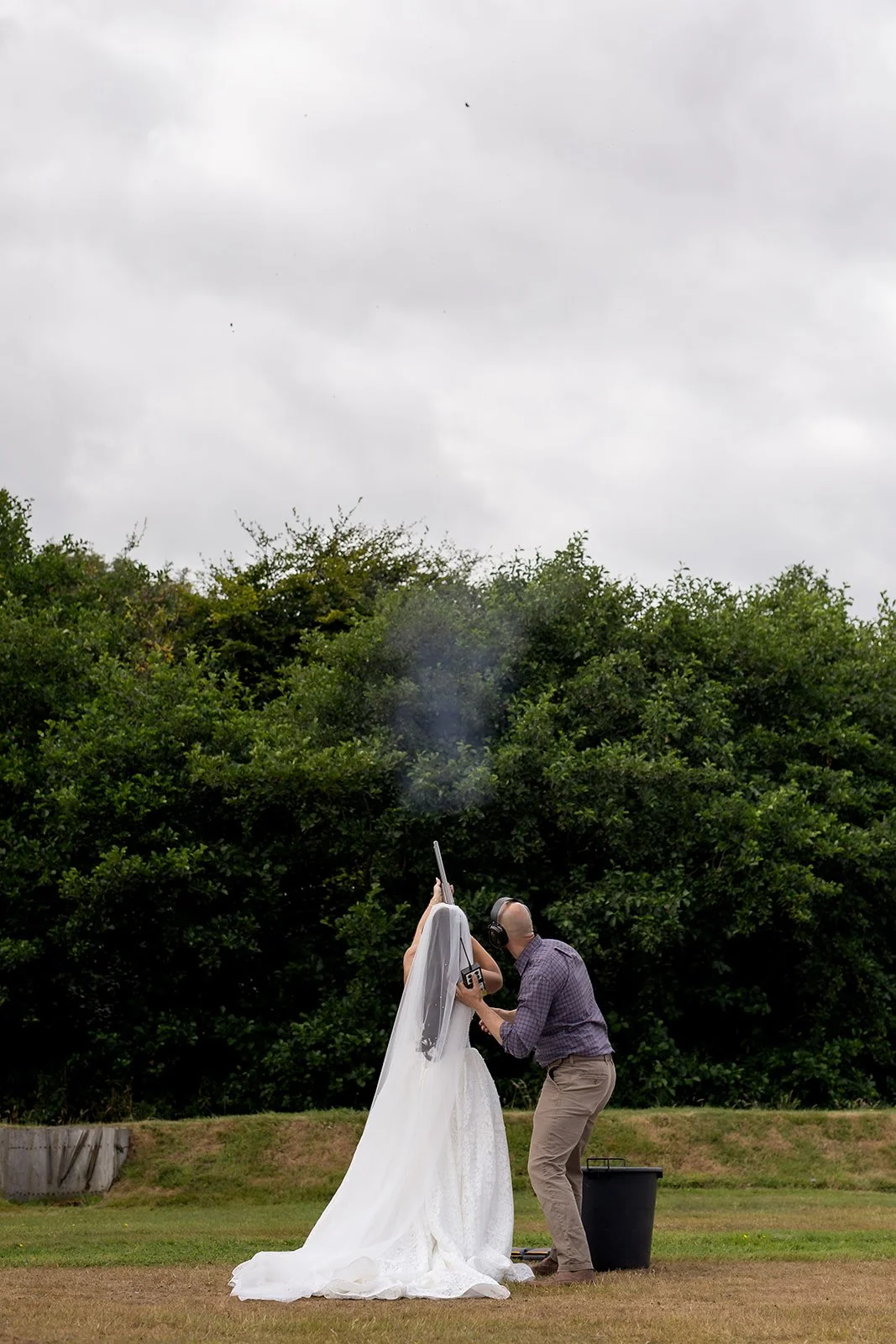 A bride in a white wedding dress and veil standing outdoors, with someone adjusting her hair or veil, under a cloudy sky with trees in the background.