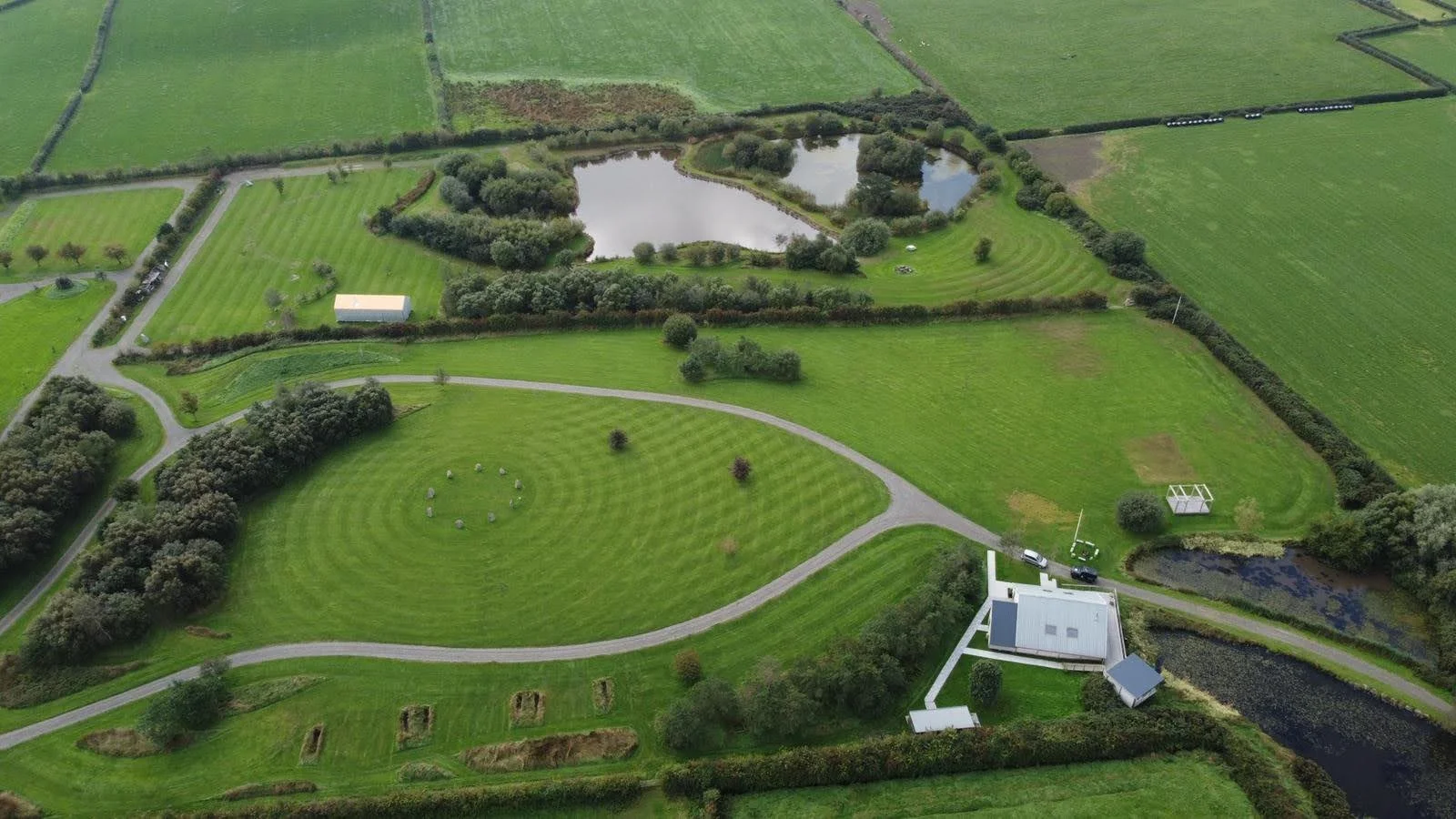Aerial view of a large grassy estate with a pond, trees, buildings, and gardens, surrounded by farmland.