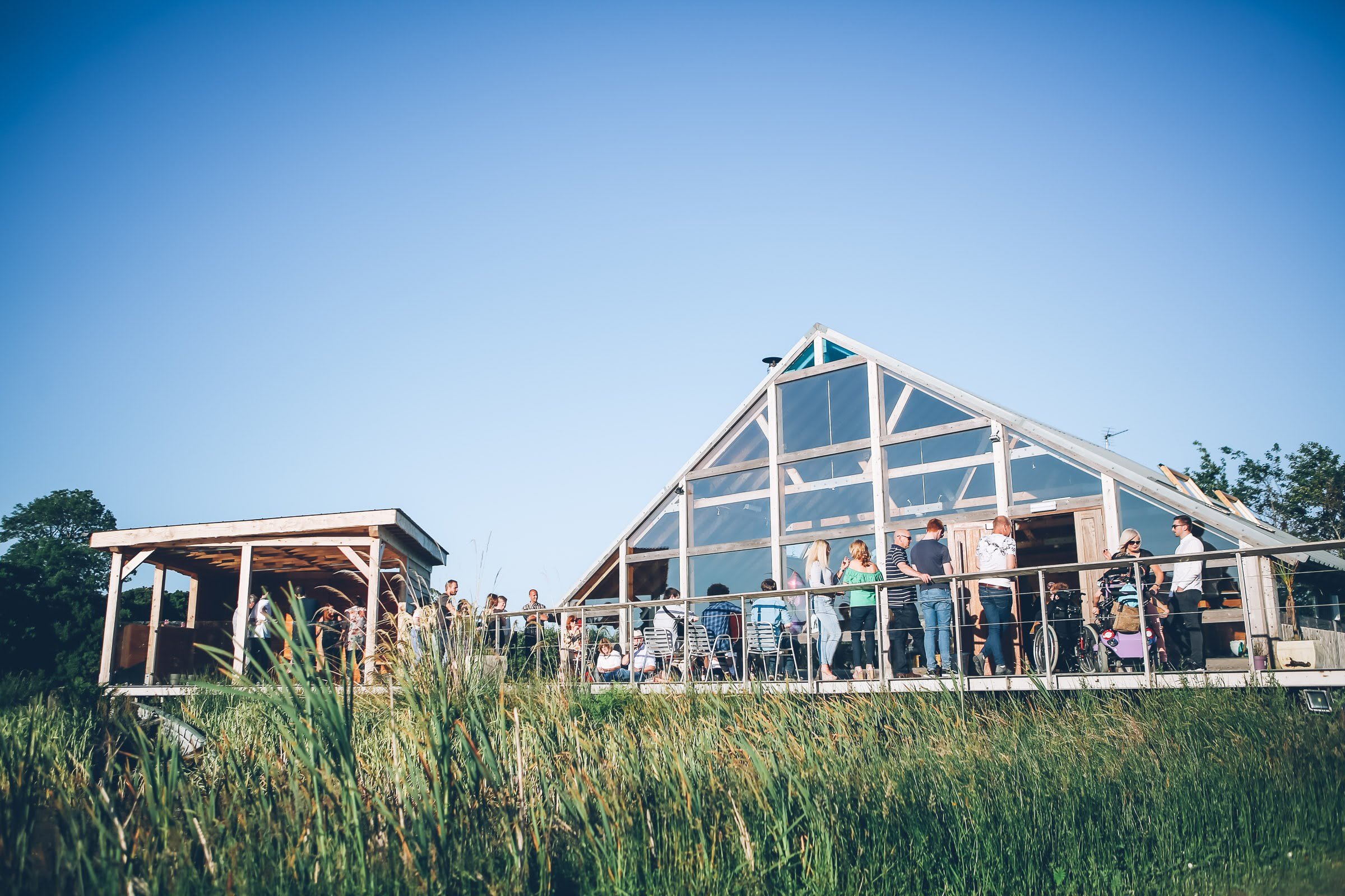 People gather around an outdoor wooden greenhouse structure with glass panels on a sunny day.