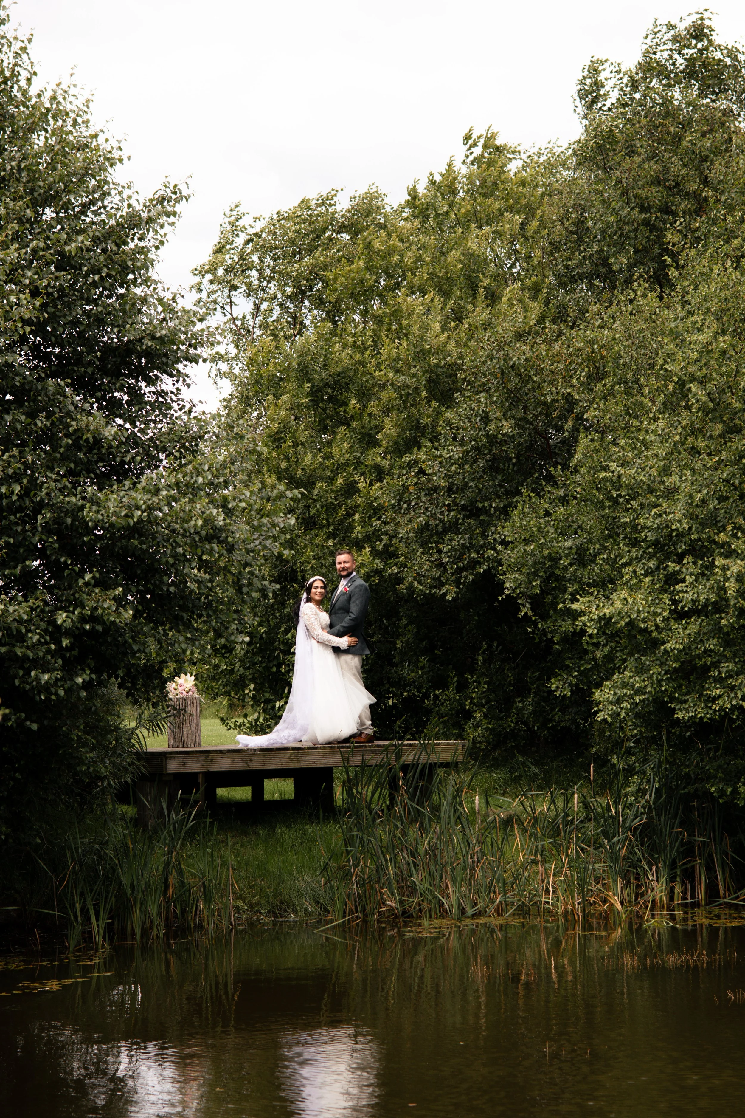 A bride and groom standing on a small wooden dock by a pond, surrounded by lush greenery and trees, with a cloudy sky overhead.