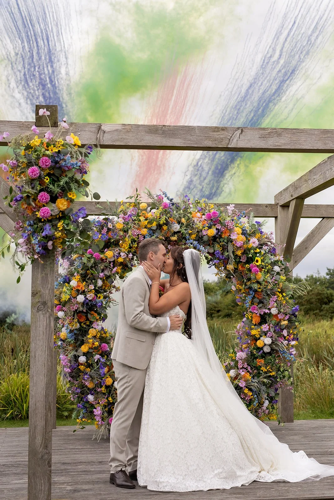 A bride and groom kiss under a floral arch on an outdoor wedding stage.