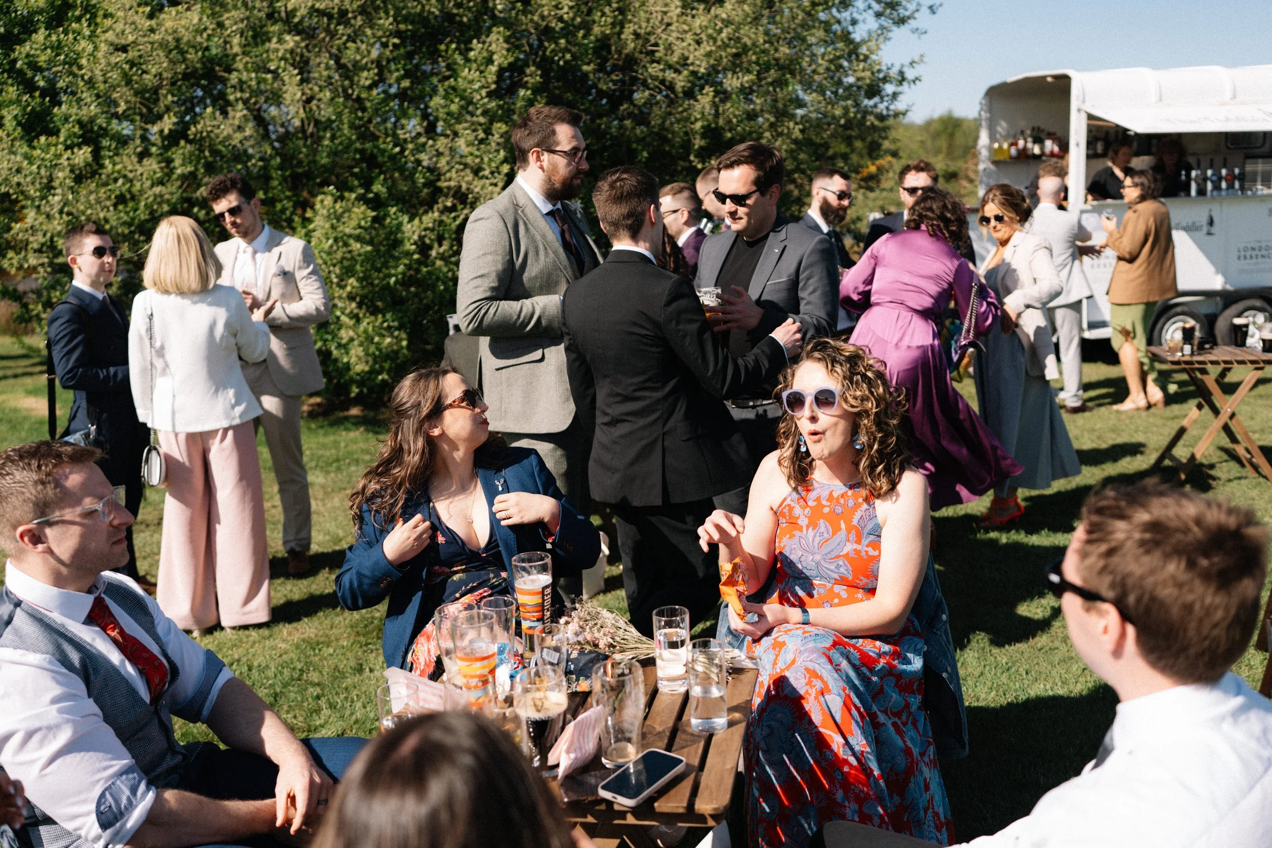 A group of people at an outdoor gathering, some are sitting at a picnic table with drinks and snacks, others are standing and chatting, with a food truck in the background.