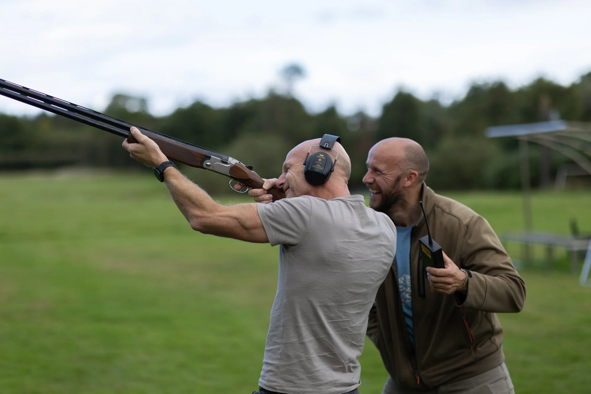 Two men outdoors, one holding and aiming a shotgun while the other assists, with a background of green grass and trees.