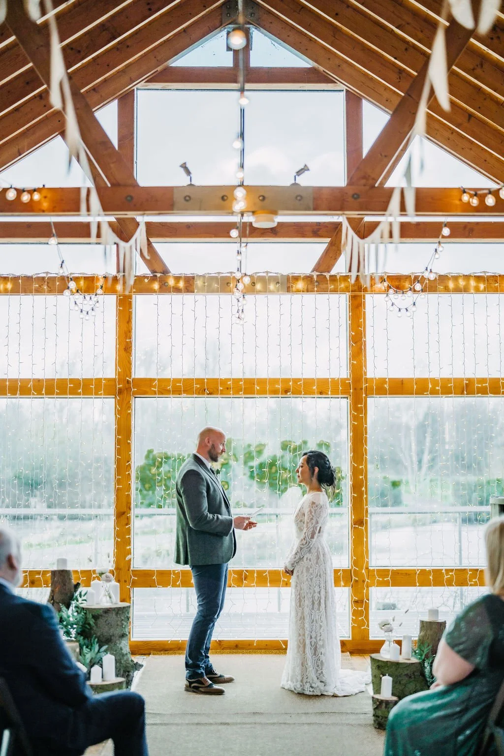 A couple getting married in a rustic indoor venue with wooden beams, string lights, and large windows, with guests seated nearby.