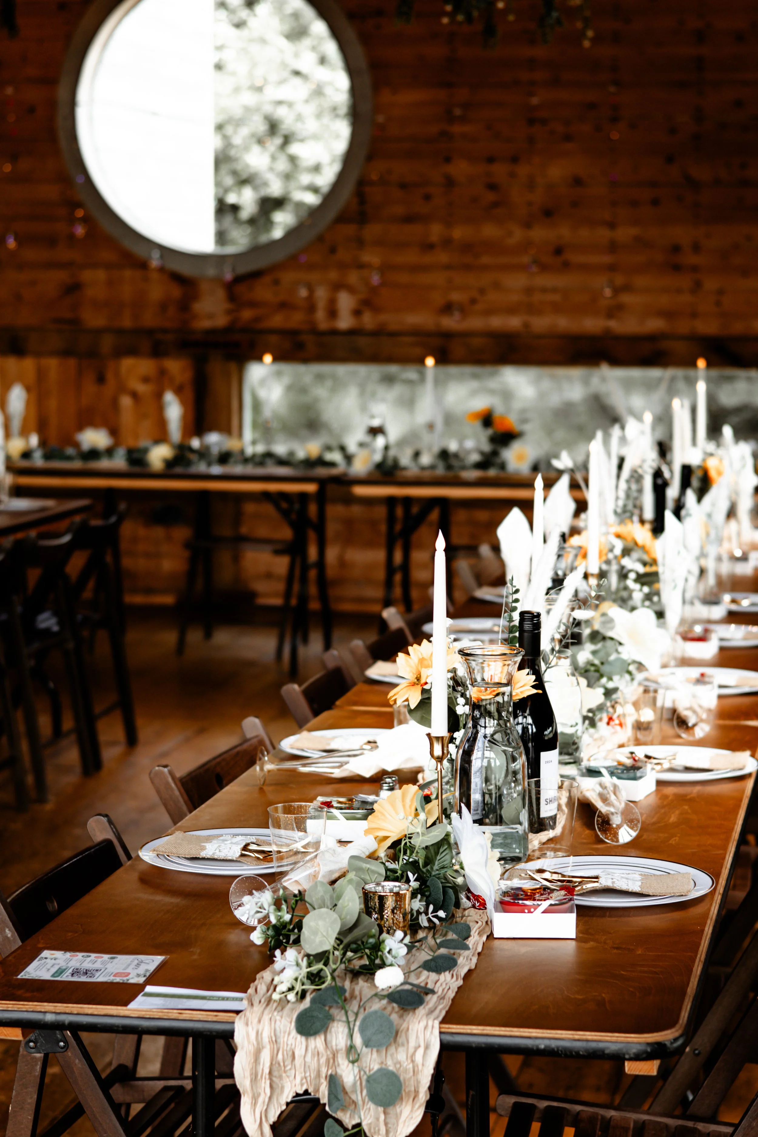 A decorated banquet table with candles, floral arrangements, and wine bottles set up inside a wooden lodge or barn with a high ceiling and large round window.