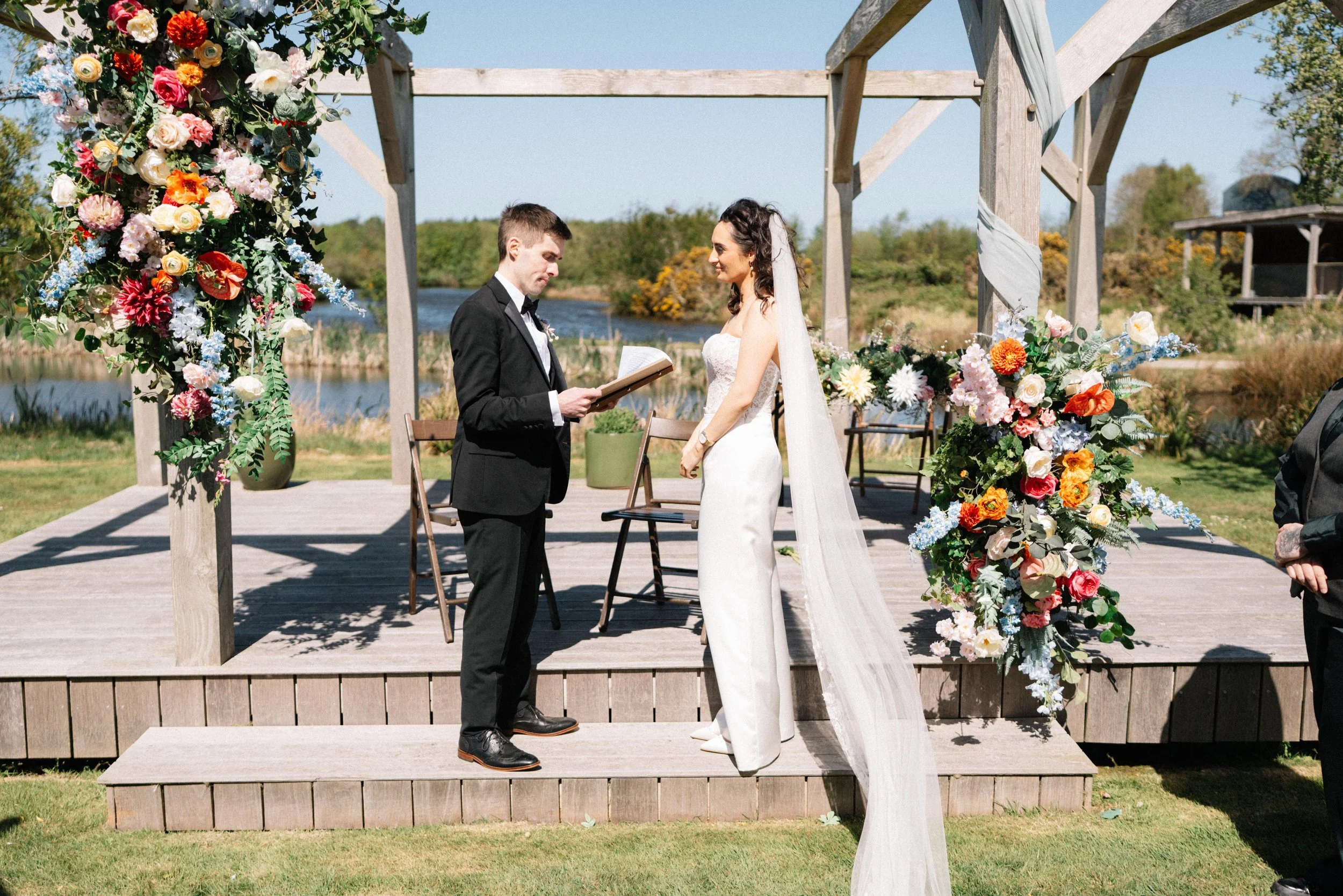 A bride and groom exchanging vows during an outdoor wedding ceremony by a lake, with floral arches and wooden framework.