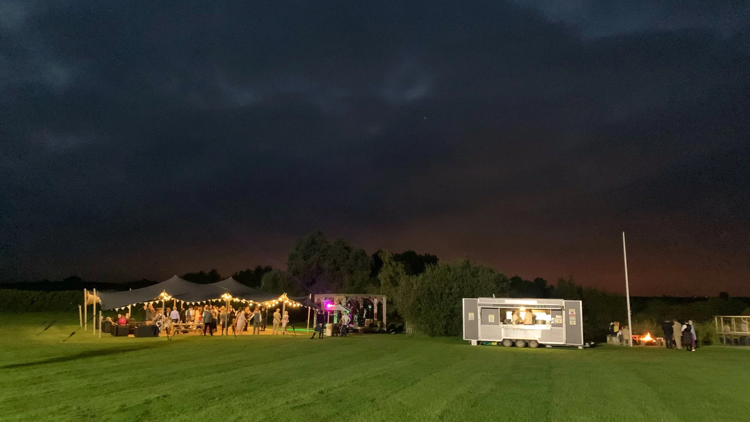 An outdoor nighttime event with a large tent, string lights, and a stage with people gathered around, on a grassy field with trees in the background and a dark sky.