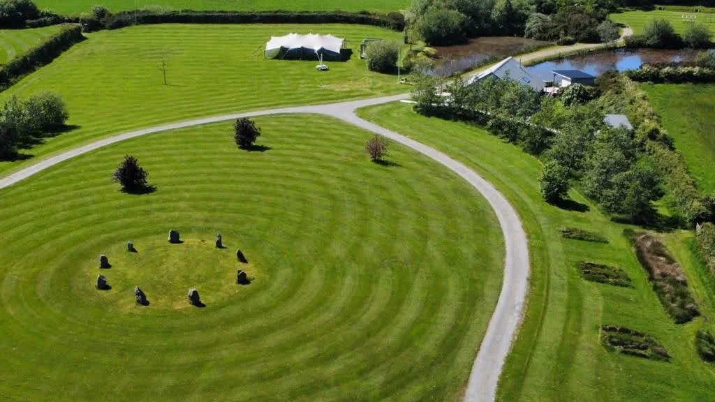 Aerial view of a large green park with concentric circular patterns and stones arranged in a circle inside the innermost circle. There are a few scattered trees, a winding path, and a building near a pond in the background.